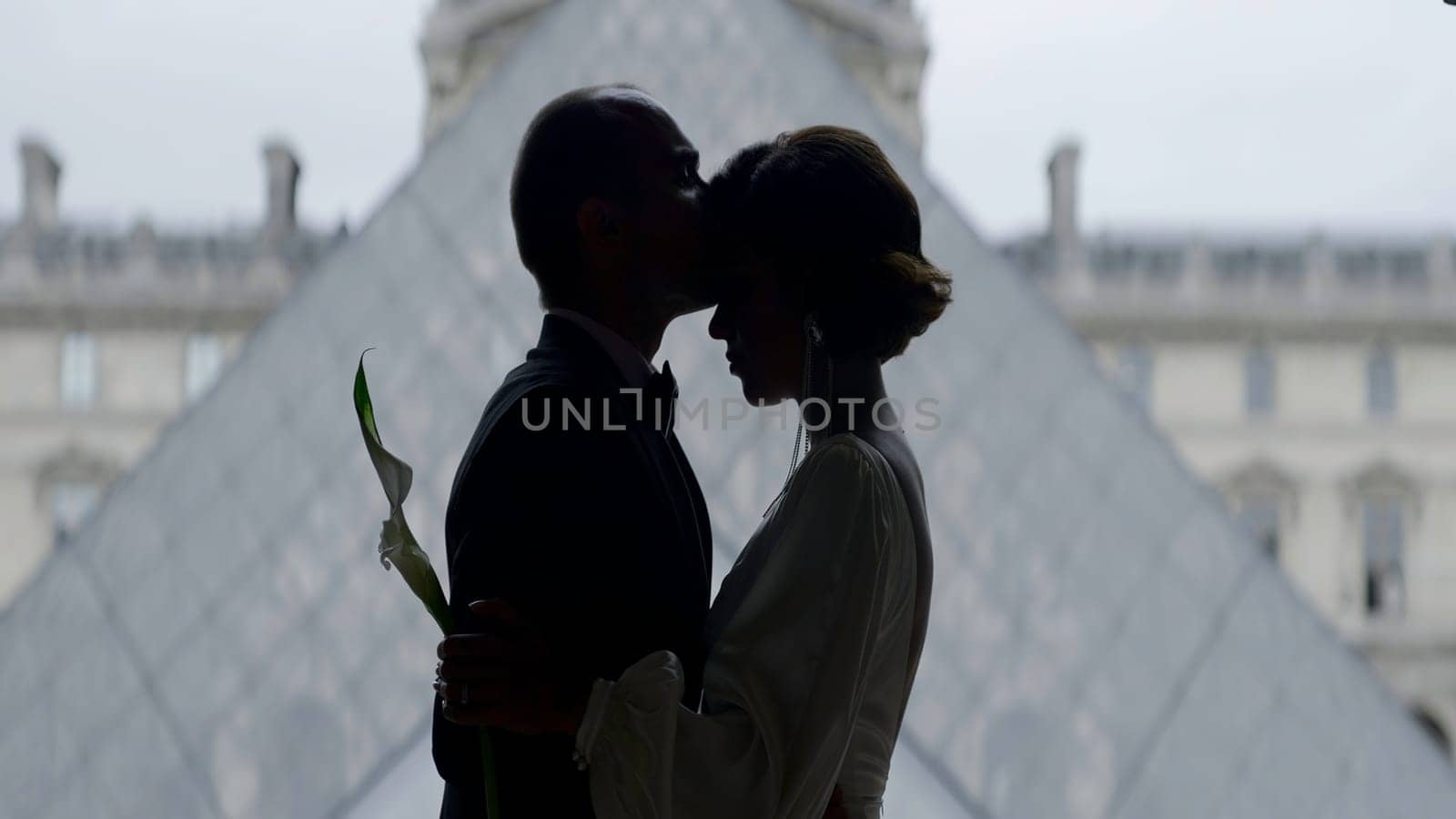 Happy couple near the Louvre museum pyramid in Paris. Action. Side view of embracing bride and groom. by Mediawhalestock