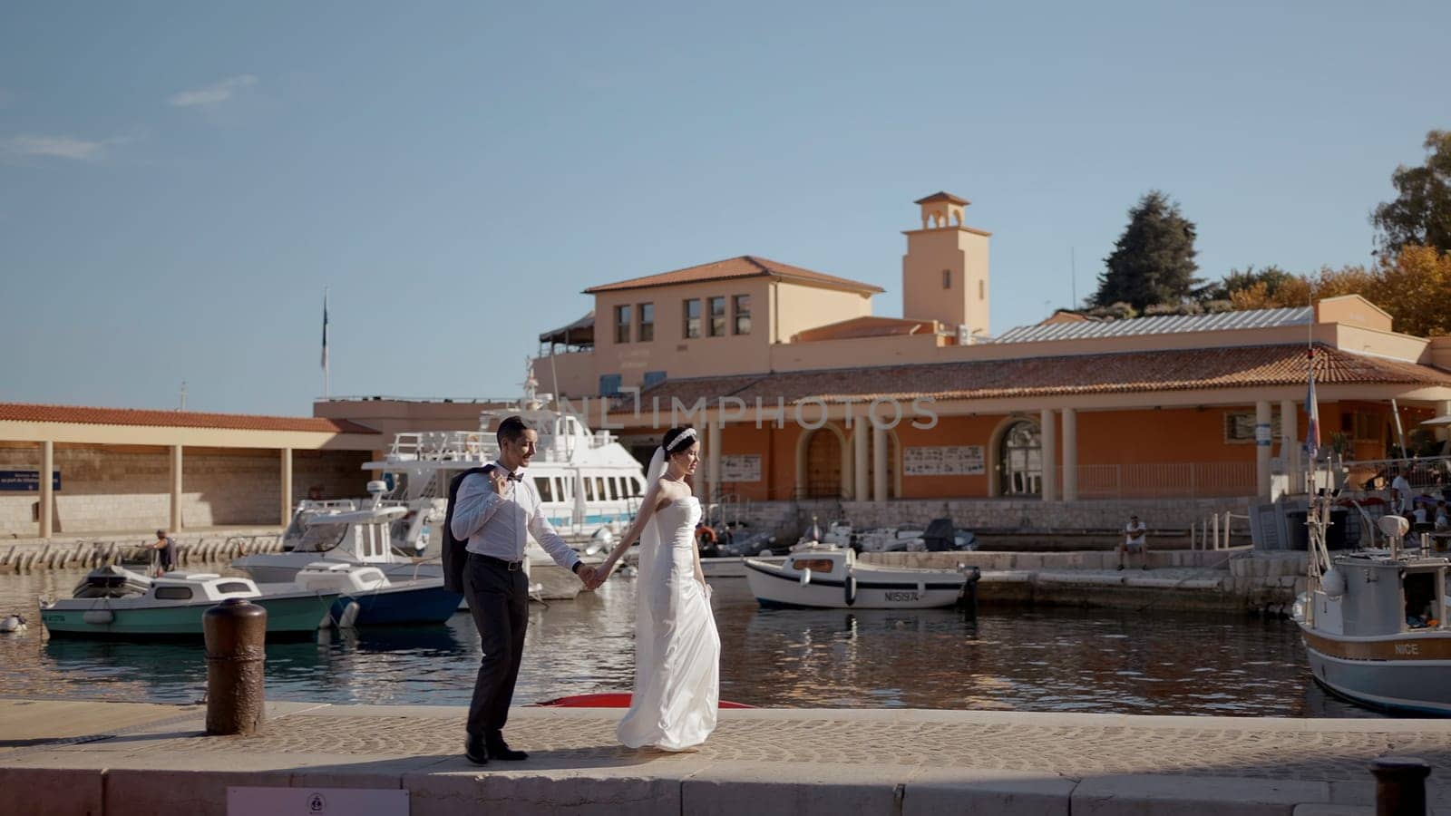 Newlyweds walking near the fountains. Action. Beautiful people together with a bride in a white tight dress and a man in tourist places near the river with parked ships. by Mediawhalestock