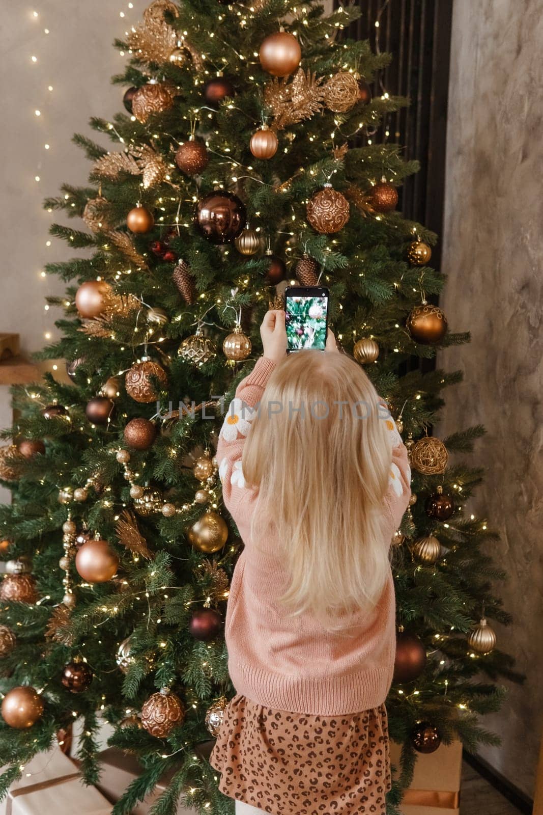 A little blonde girl photographs balloons on a Christmas tree in a festive interior decorated in a New Year's style. The concept of a merry Christmas