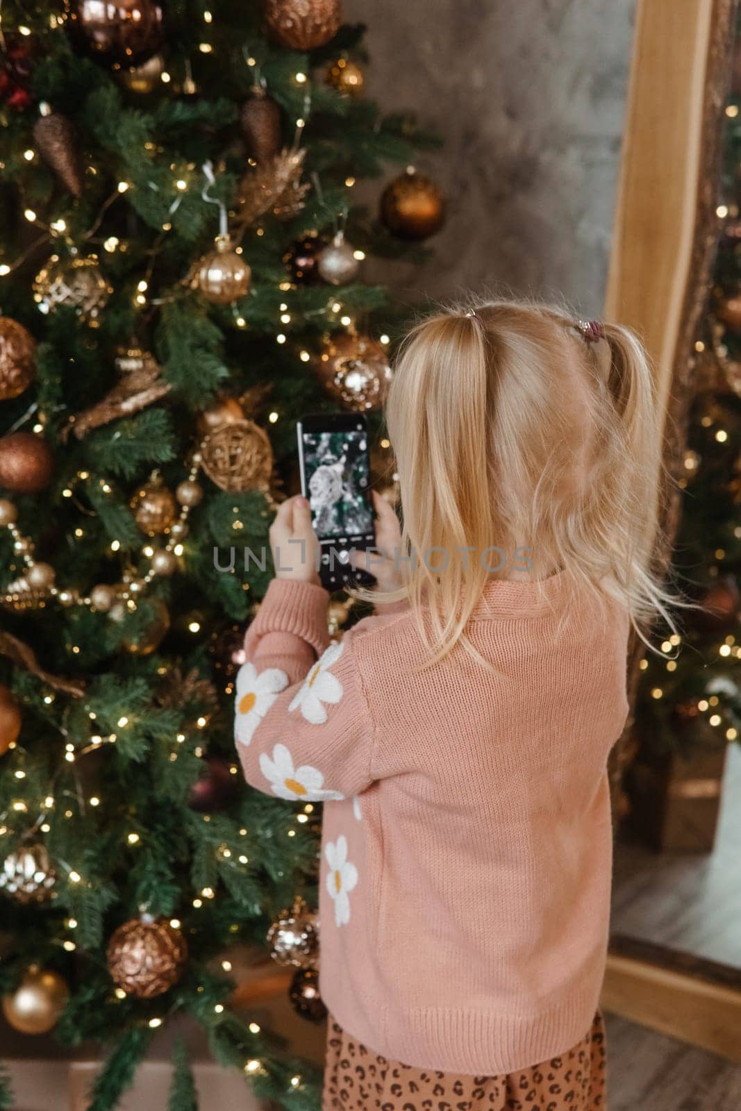 A little blonde girl photographs balloons on a Christmas tree in a festive interior decorated in a New Year's style. The concept of a merry Christmas