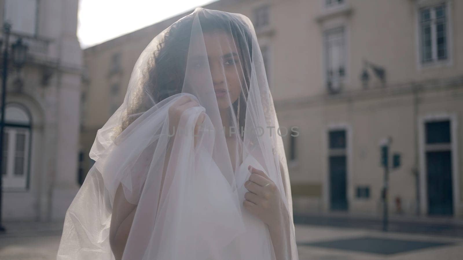 Beautiful woman with white veil in city. Action. Bride in white veil poses in old town. Woman with veil on city street on sunny summer day by Mediawhalestock