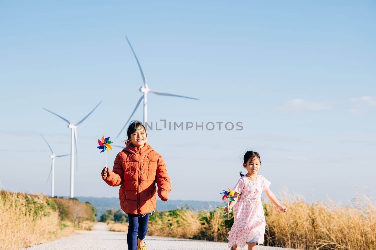 Two children girl with pinwheel happily near windmills. Family moments at turbines symbolize clean energy. Children joy windmill circle in sky represents sustainable cheerful community.