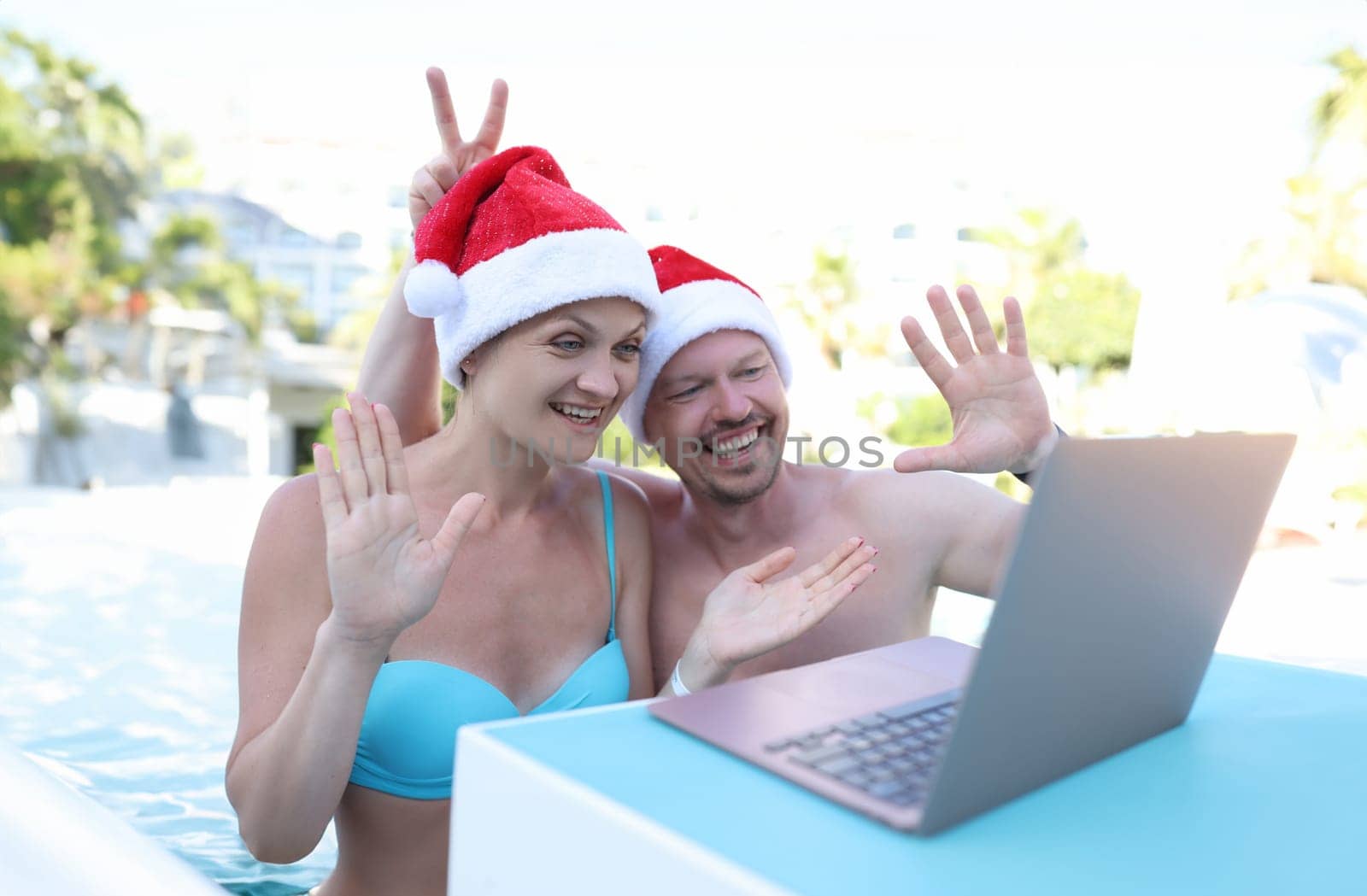 Man and woman in santa hats waving hand at laptop screen on vacation at resort by kuprevich