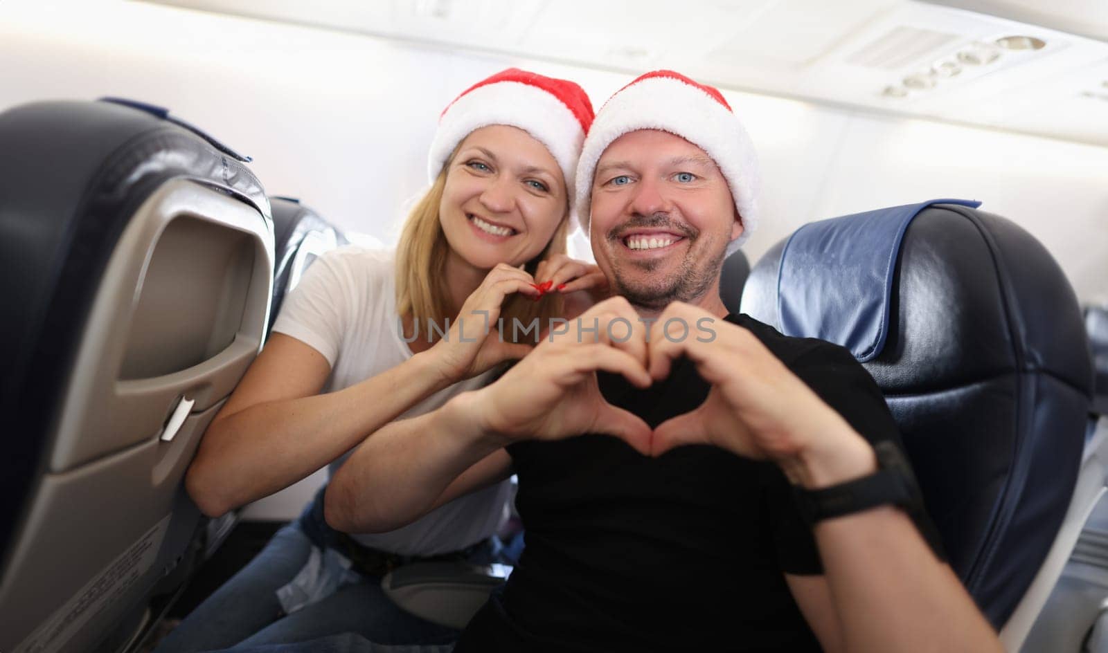 Man and woman in santa hats flying in airplane and showing heart with hands by kuprevich