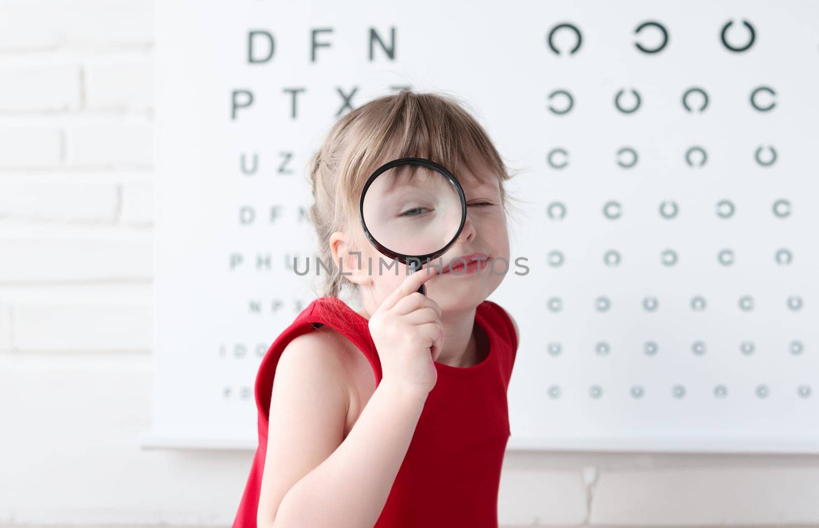 Little girl with magnifying glass standing on background of table for eye test. Eye examination in children concept