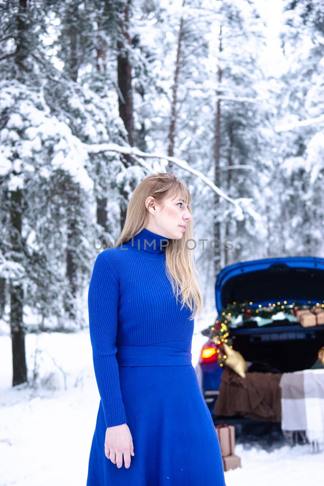 Woman in winter snowy forest in blue dress next to blue car decorated with Christmas decor. Christmas and winter holidays concept. by Annu1tochka