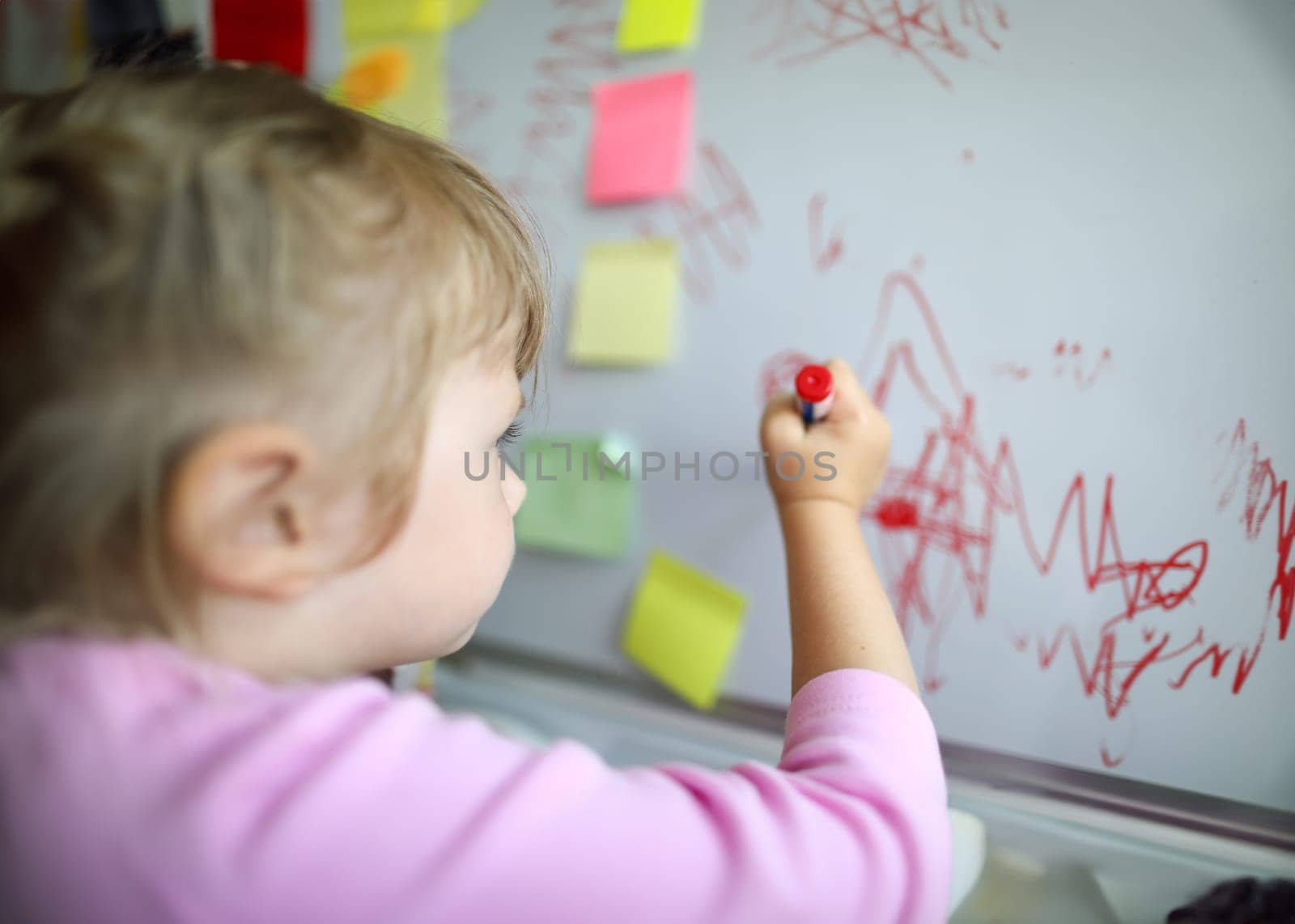 Focus on tender small hand of little girl holding big red marker and drawing cute childish paintings on glass board in modern classroom. Creative childhood concept