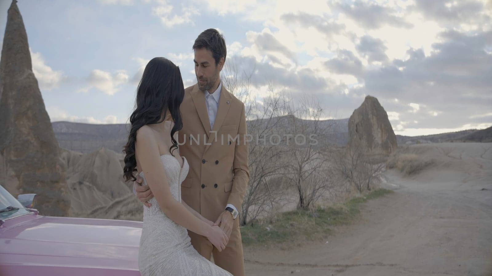 Beautiful couple with car on background of mountains. Action. Elegant outfits of newlyweds vacationing in mountains. Stylish newlyweds sit on hood of car on background of canyon.