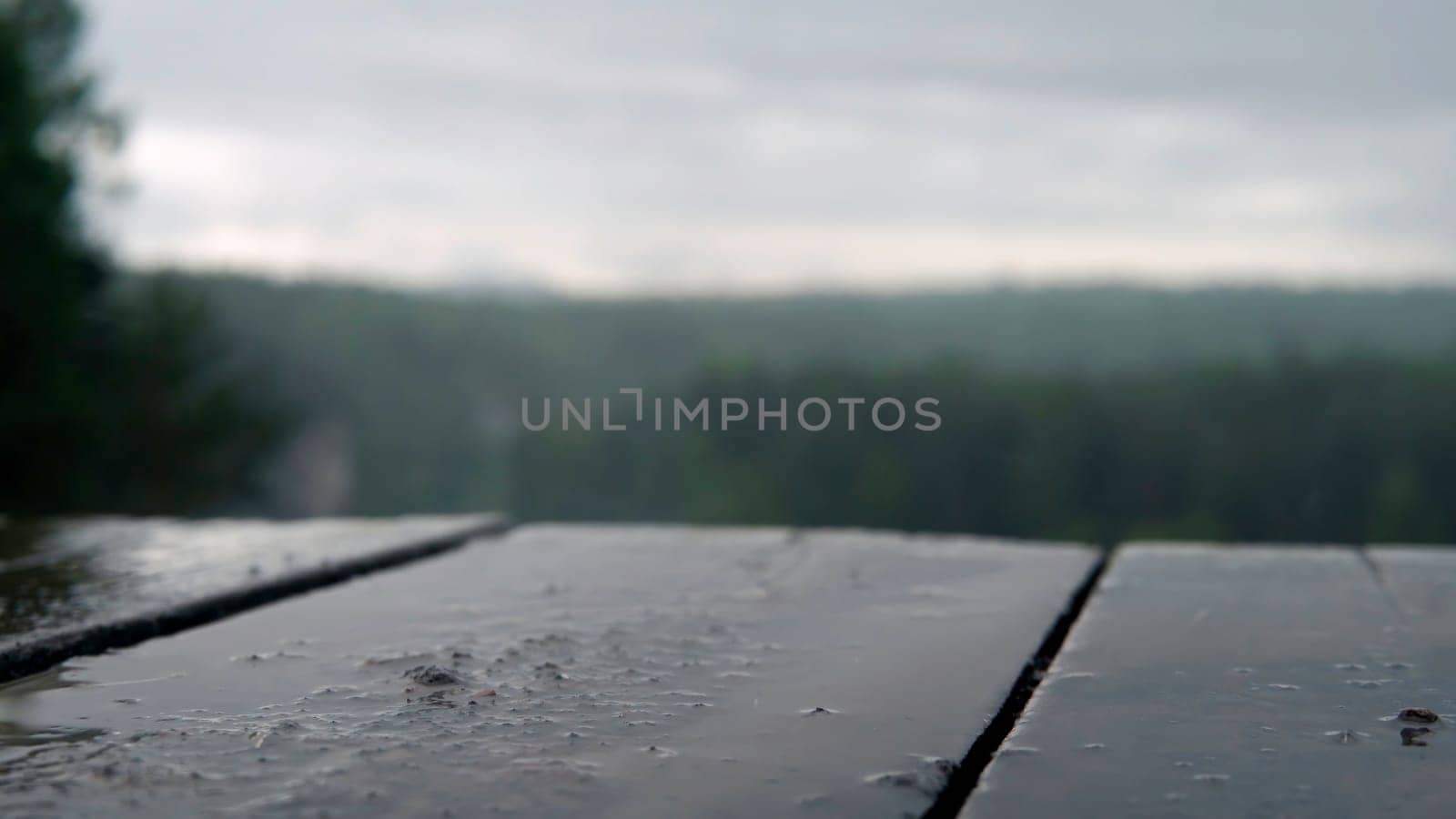 Raindrops on wooden table on background of green forest. Stock footage. Cloudy rainy weather hung over park and rain drops hit picnic table.