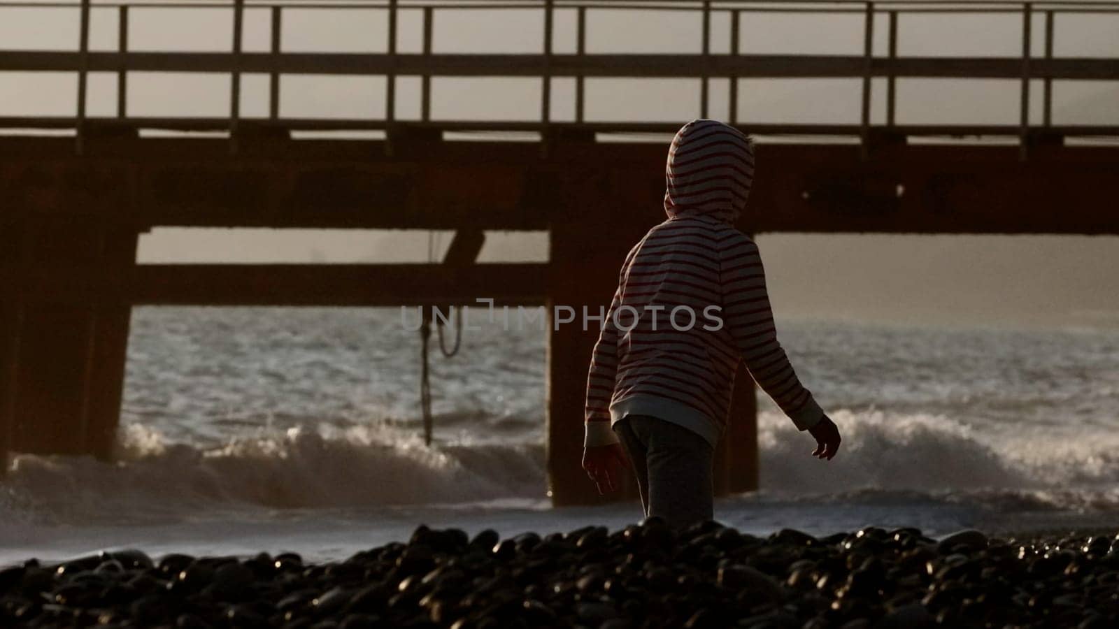 Children walking along the seashore. Creative.Little boys running along the stone beach from the waves next to buoys and bridges and you can see the sunny sky. by Mediawhalestock