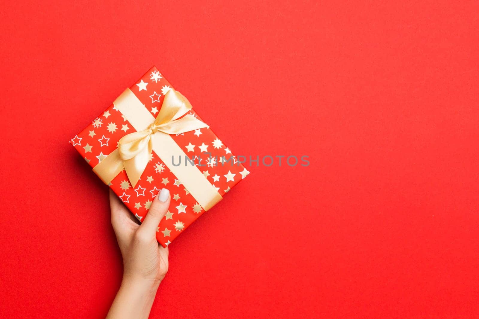 Female's hands holding striped gift box with colored ribbon on red background. Christmas concept or other holiday handmade present box, concept top view with copy space.