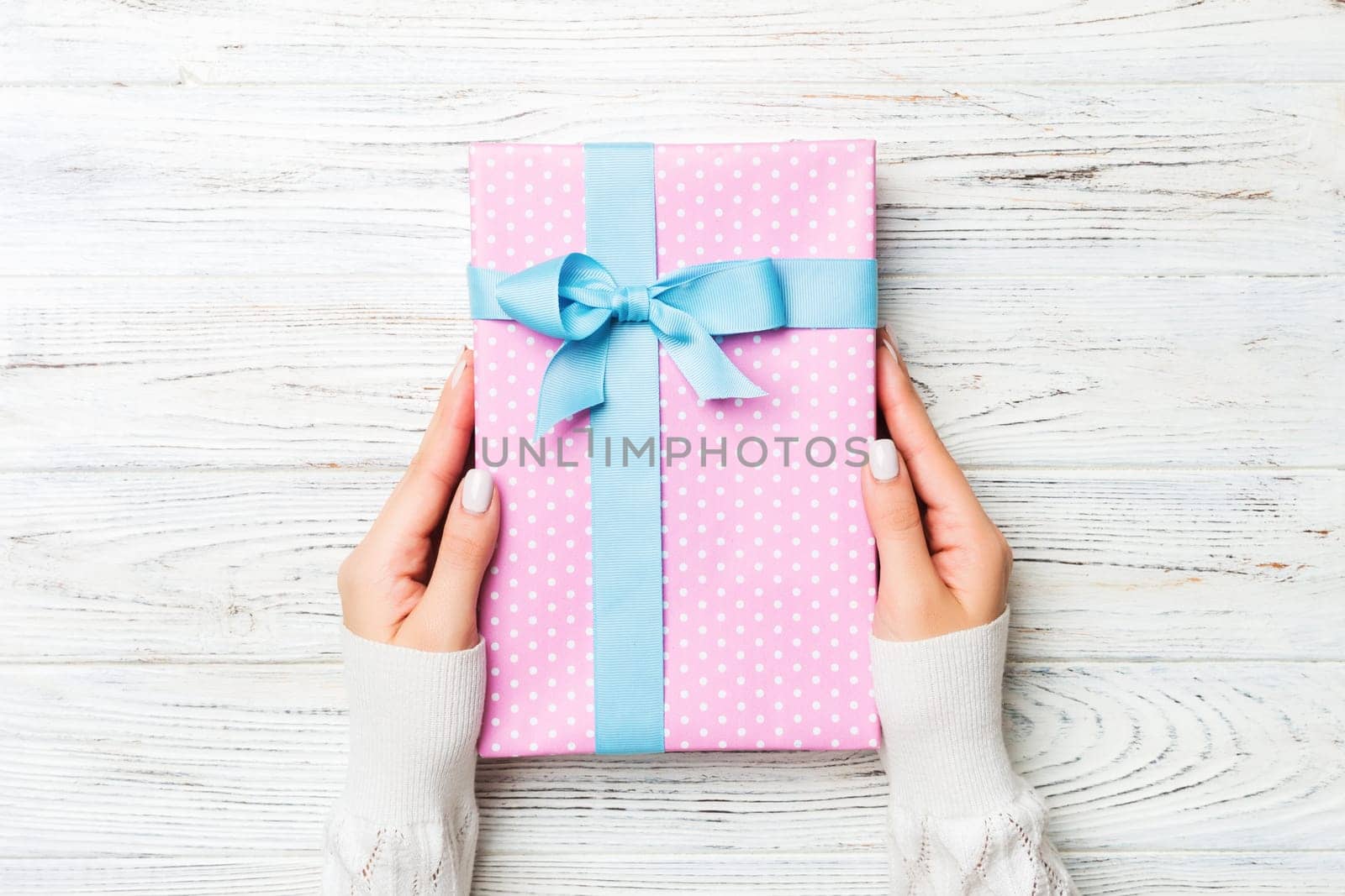 Top view woman hands holding present box with colored bow on white rustic wooden background with copy space.