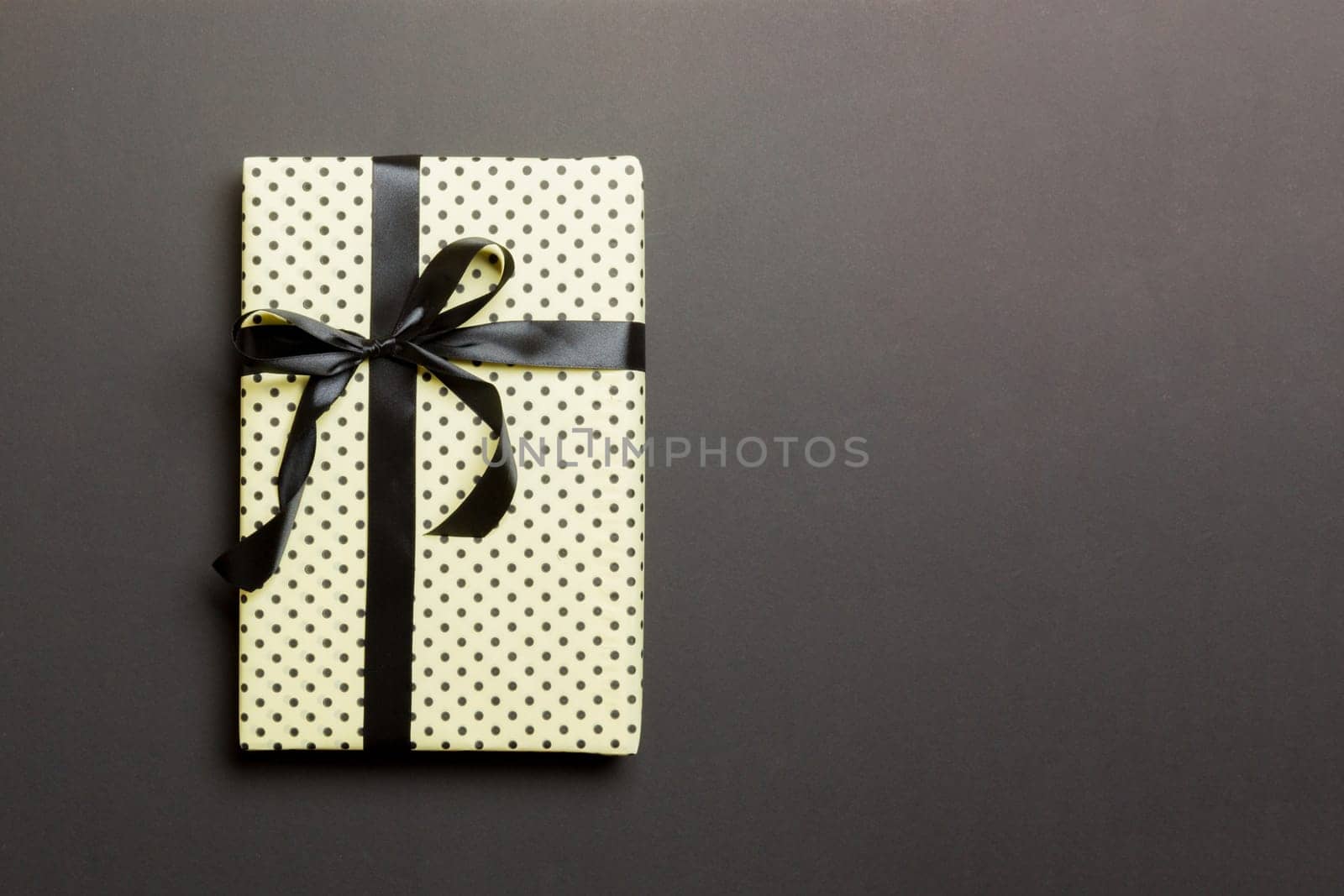 wrapped Christmas or other holiday handmade present in paper with black ribbon on black background. Present box, decoration of gift on colored table, top view with copy space.