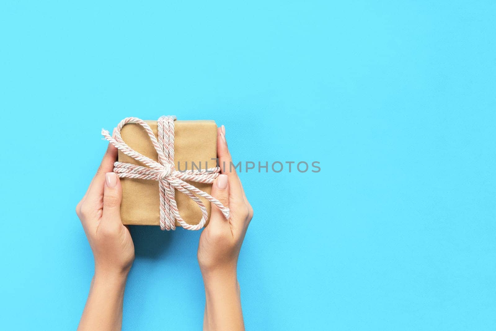 Woman hands give wrapped valentine or other holiday handmade present in paper with blue ribbon. Present box, decoration of gift on table, top view with copy space.