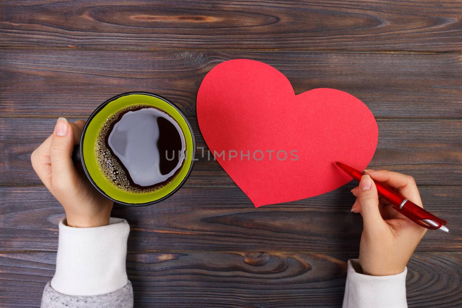 Hands holding a cup of coffee and writing valentine. valentines day decorations on the table. A rustic wooden valentine background. Top view. Copy space.
