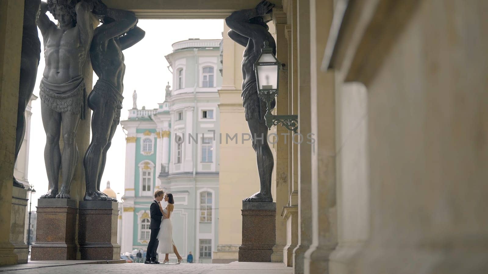 Beautiful newlyweds hug at old building. Action. Small silhouette of newlyweds in distance under arch of tall statues. Beautiful shot with tall statues under arch of building and newlyweds.