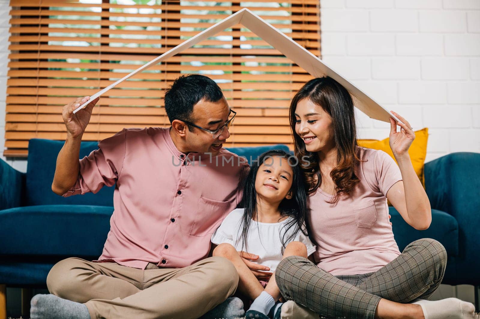 A portrait of a harmonious family sitting on a couch holding a cardboard roof radiating happiness and security in their new house. Asian parents and their daughter are all smiles.