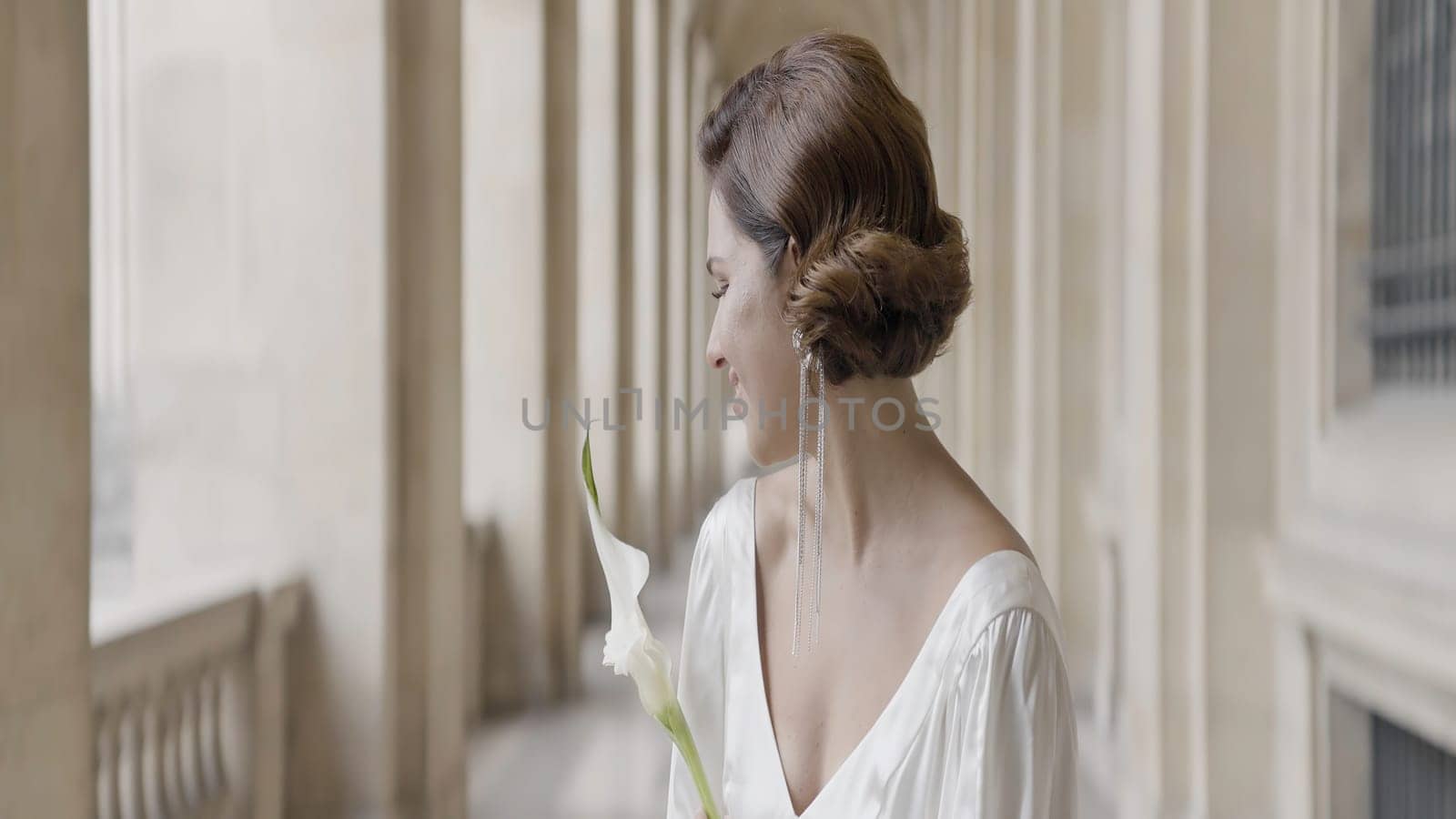 A beautiful model in a wedding dress and a white flower branch. Action. A young woman gently waving a long dress in different directions standing next to the architectural walls of the building. by Mediawhalestock