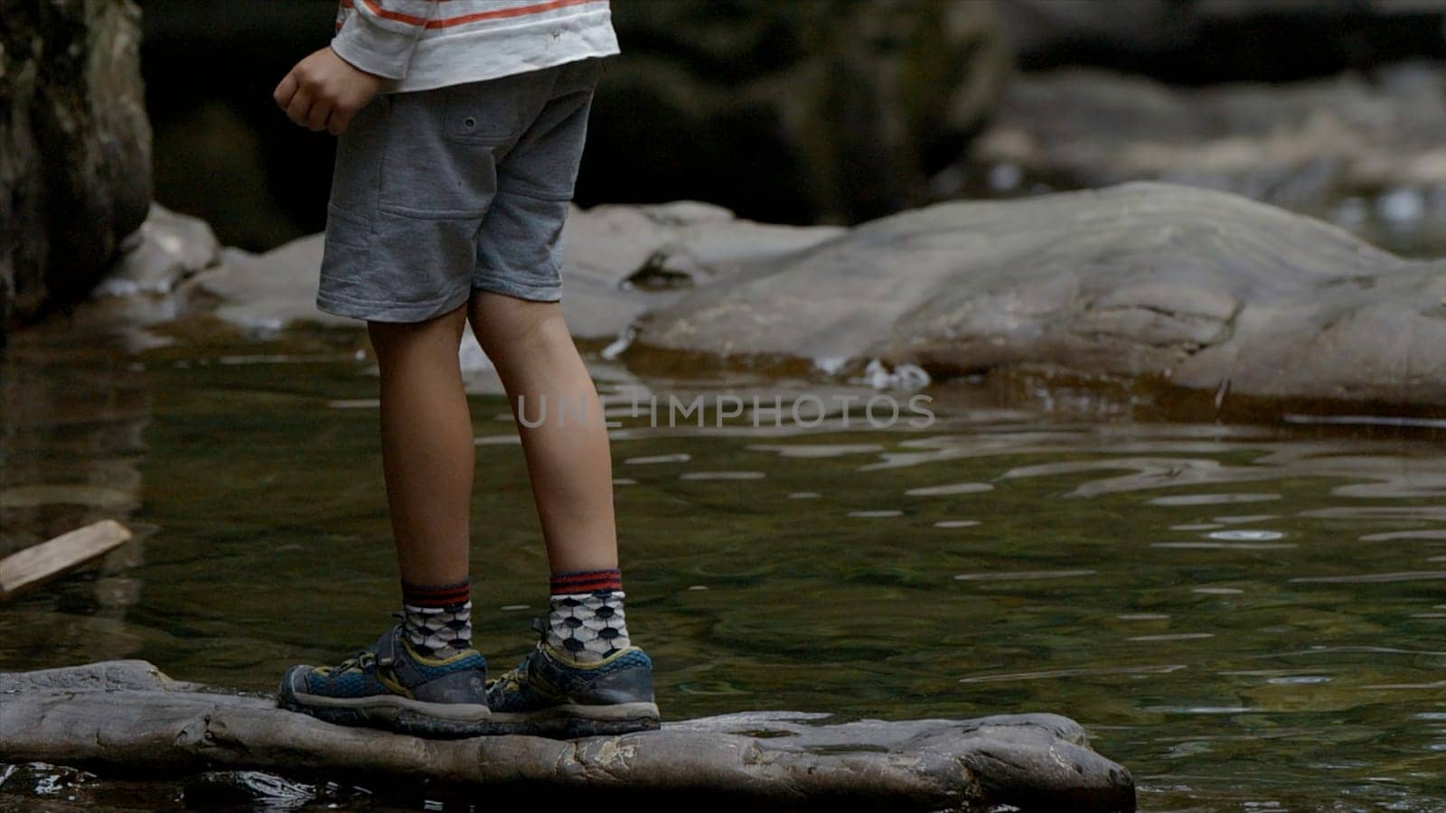 Legs of a teenager young boy in sportswear on a rock above river. Creative. Boy on a stone above rippling water surface. by Mediawhalestock