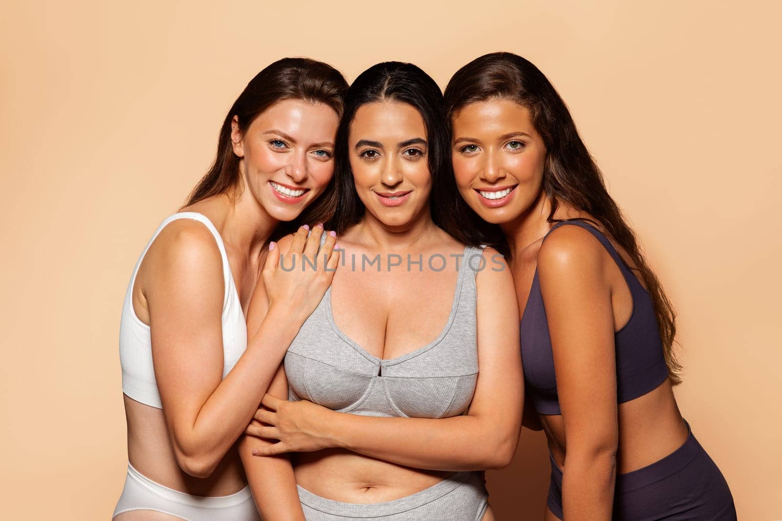 Three cheerful arab, european, latin women in underwear embracing and smiling at camera, enjoy body positive on warm background. Friendship, health, beauty care, fit and weight loss