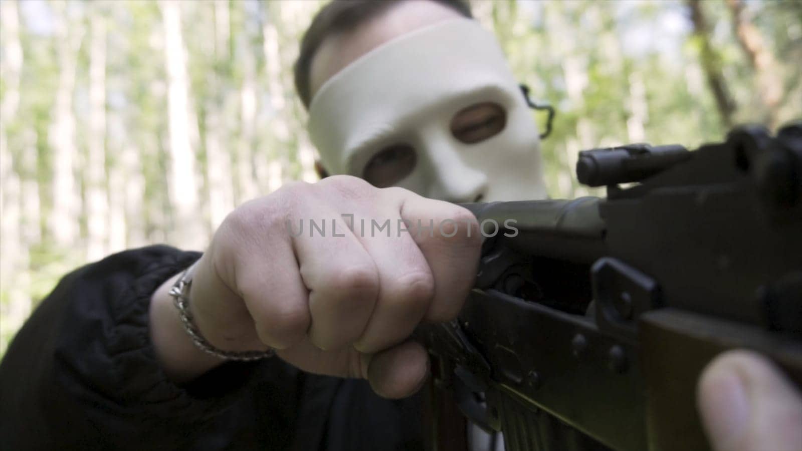 Close up of a man wearing white mask, holding rifle on green forest background, weapon and war concept. A militant loading a cartridge into the chamber of a gun, slow motion.