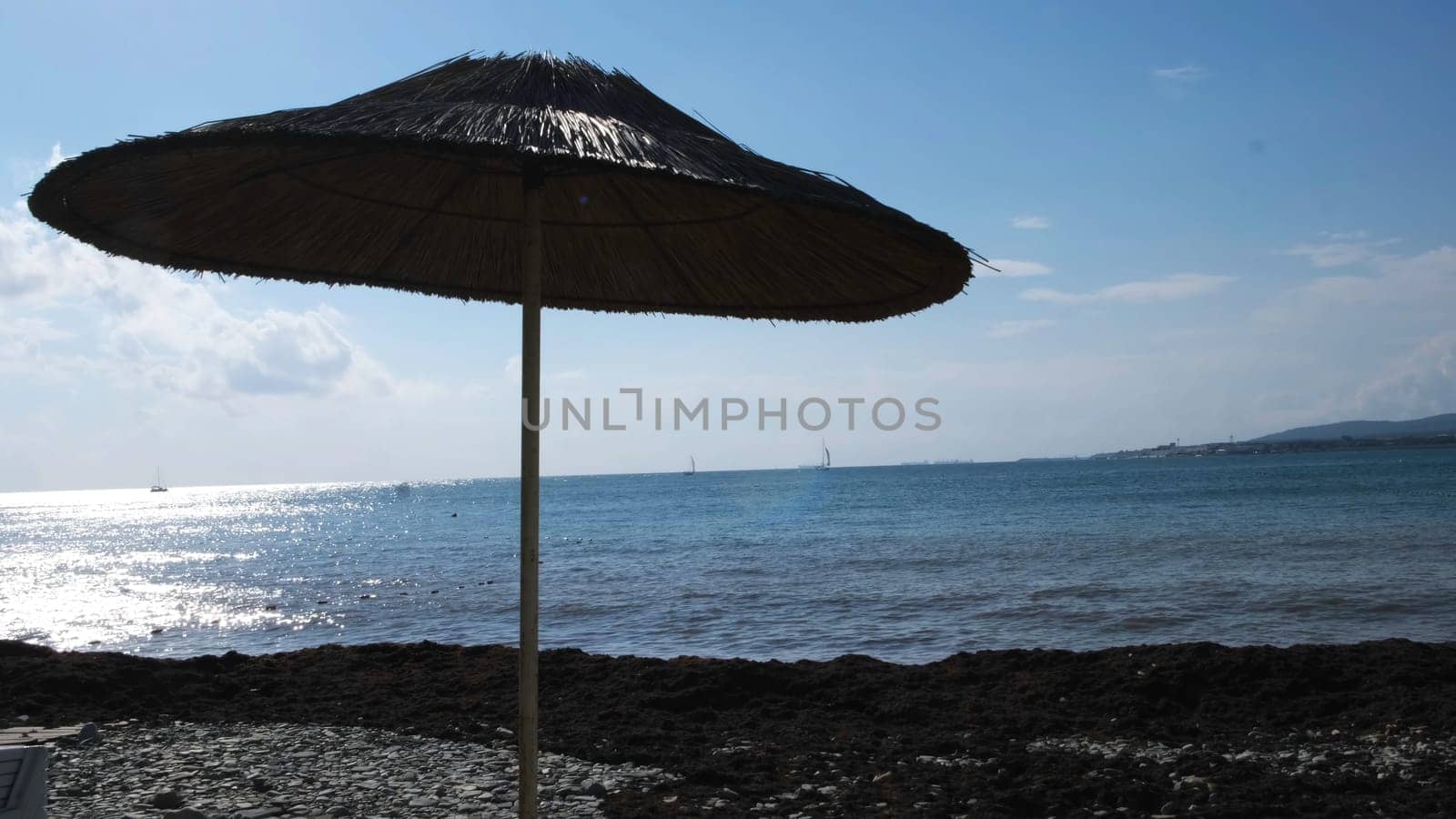 A straw umbrella on a beautiful pebble beach. Summer straw umbrella and the reflection of the sun in rippled water surface, summer holiday concept.