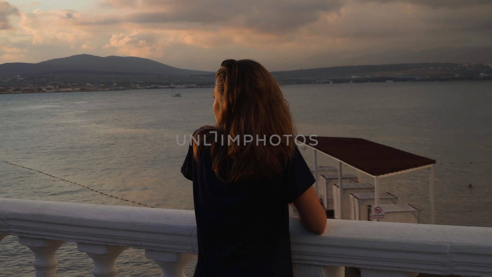 Woman on embankment at sunset. Media. Young woman stands at railing on background seascape at sunset after rain. Beautiful landscape opens up to attractive woman on embankment.