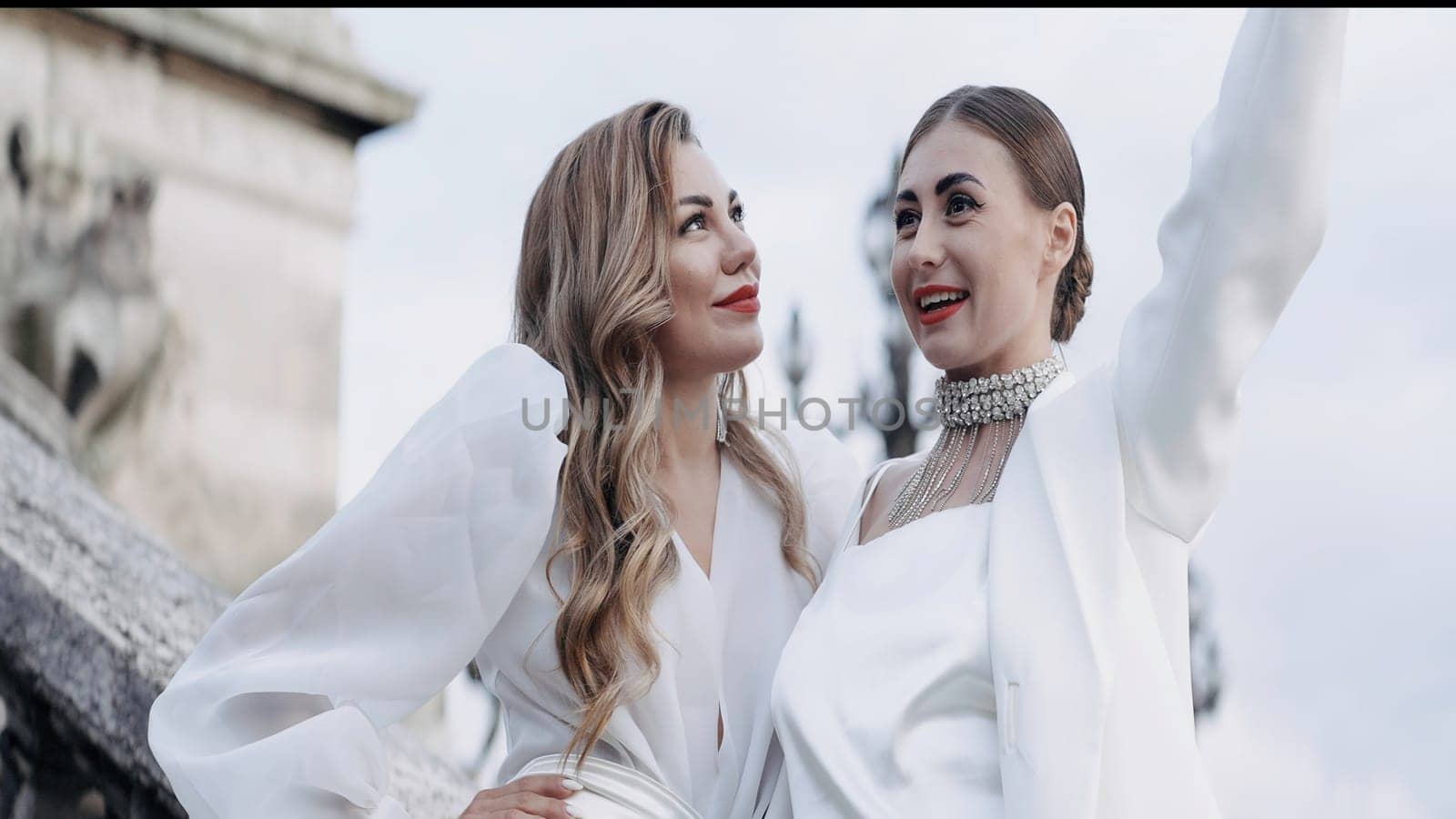 Two beautiful women blondes with red lips posing outdoors. Action. Adult ladies in white clothes holding a hat made of feathers, concept of fashion