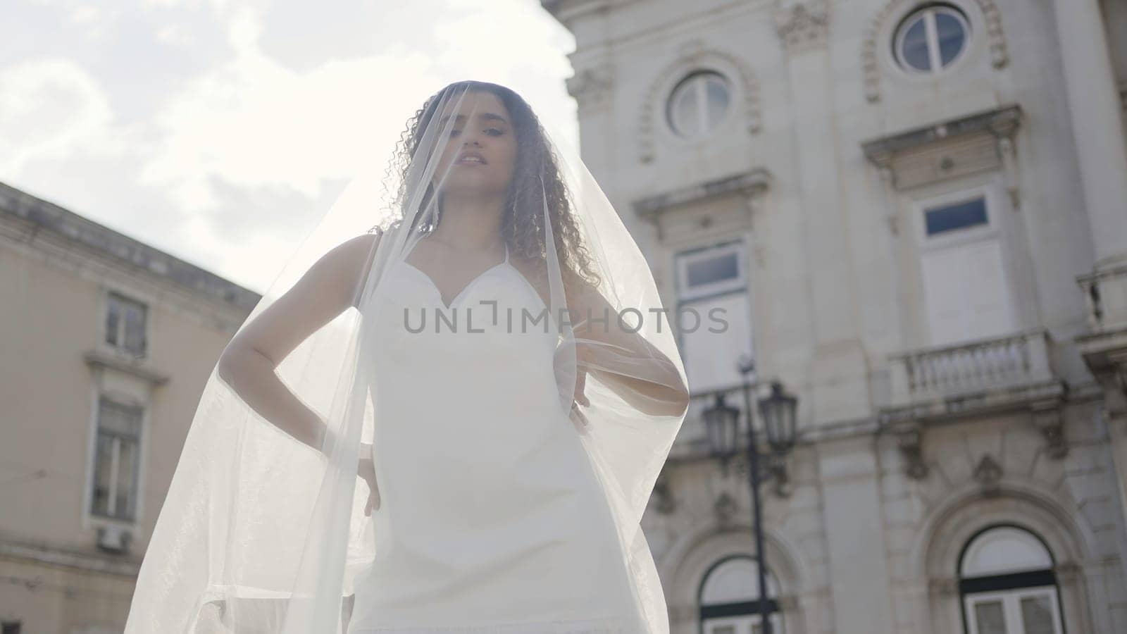 Elegant bride in short white dress and long veil. Action. Summer photosession in the old city street of a young woman with curly hair. by Mediawhalestock