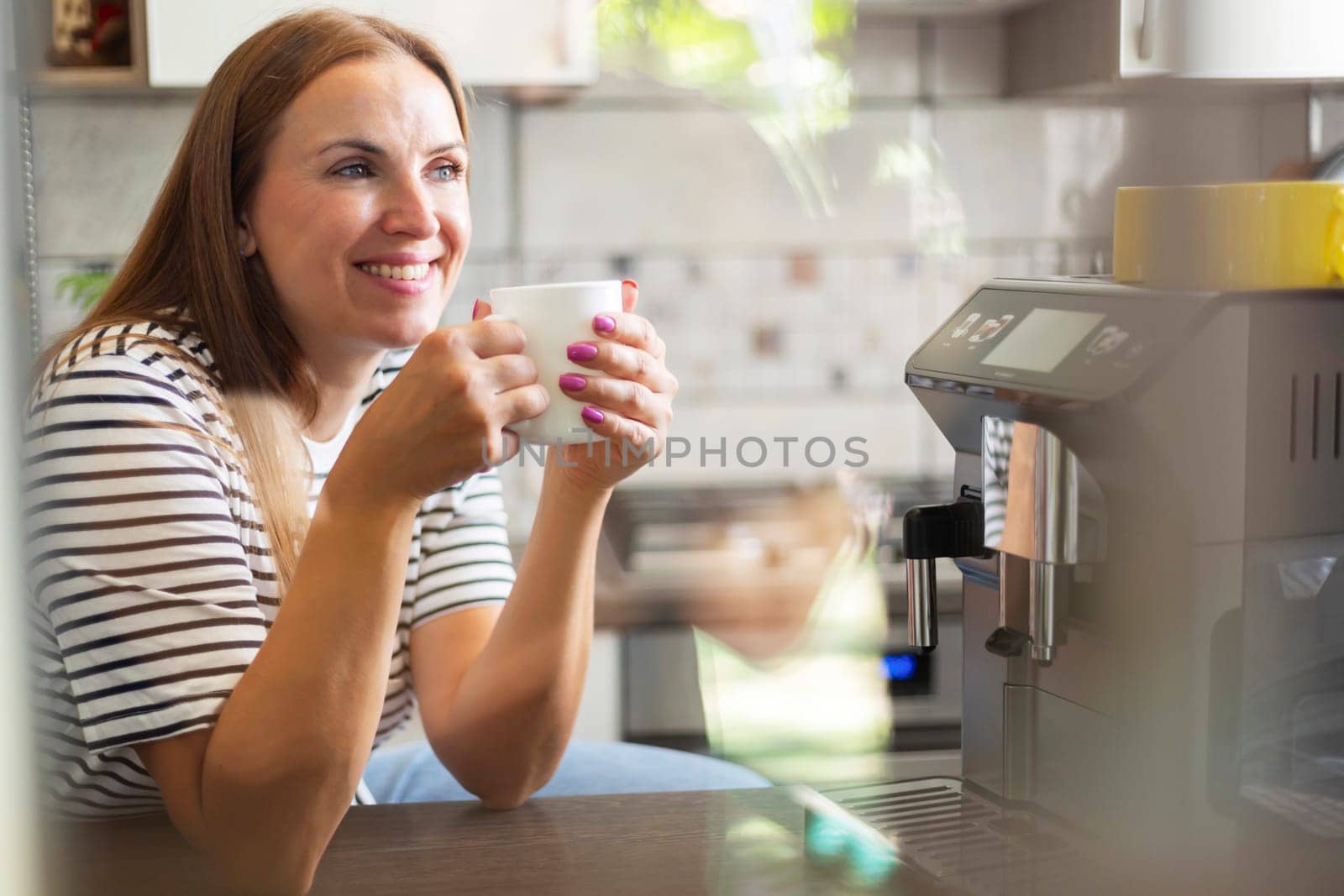 Young woman enjoying freshly prepared aromatic coffee near modern coffee machine in kitchen.