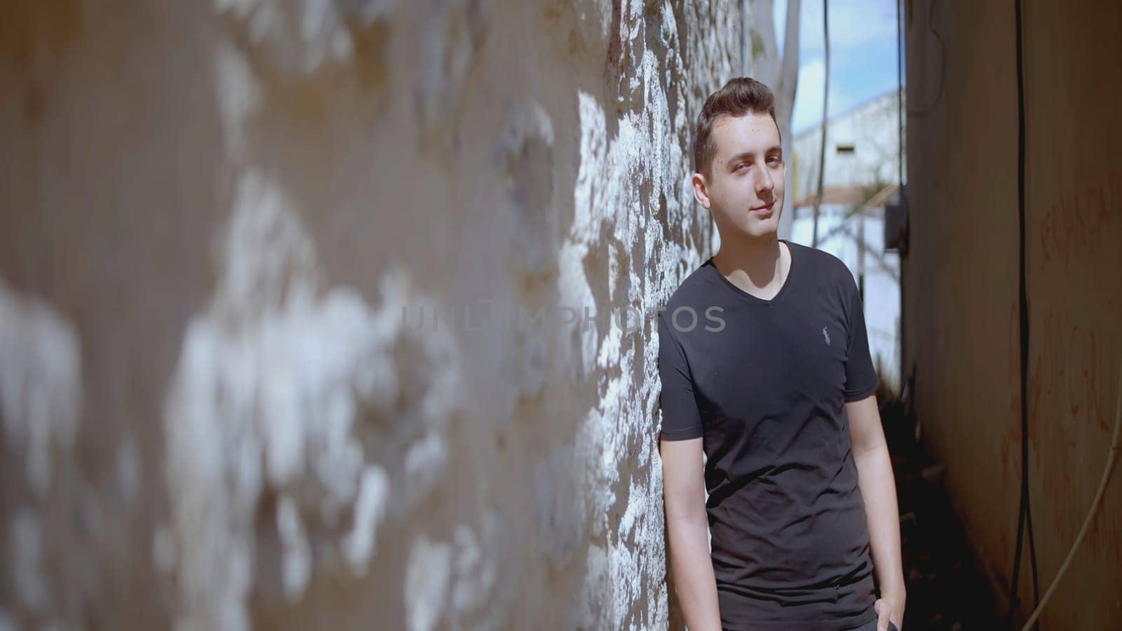 Attractive young man is standing against wall. Action. Man poses leaning against wall. Handsome man stands by stone wall on street by Mediawhalestock