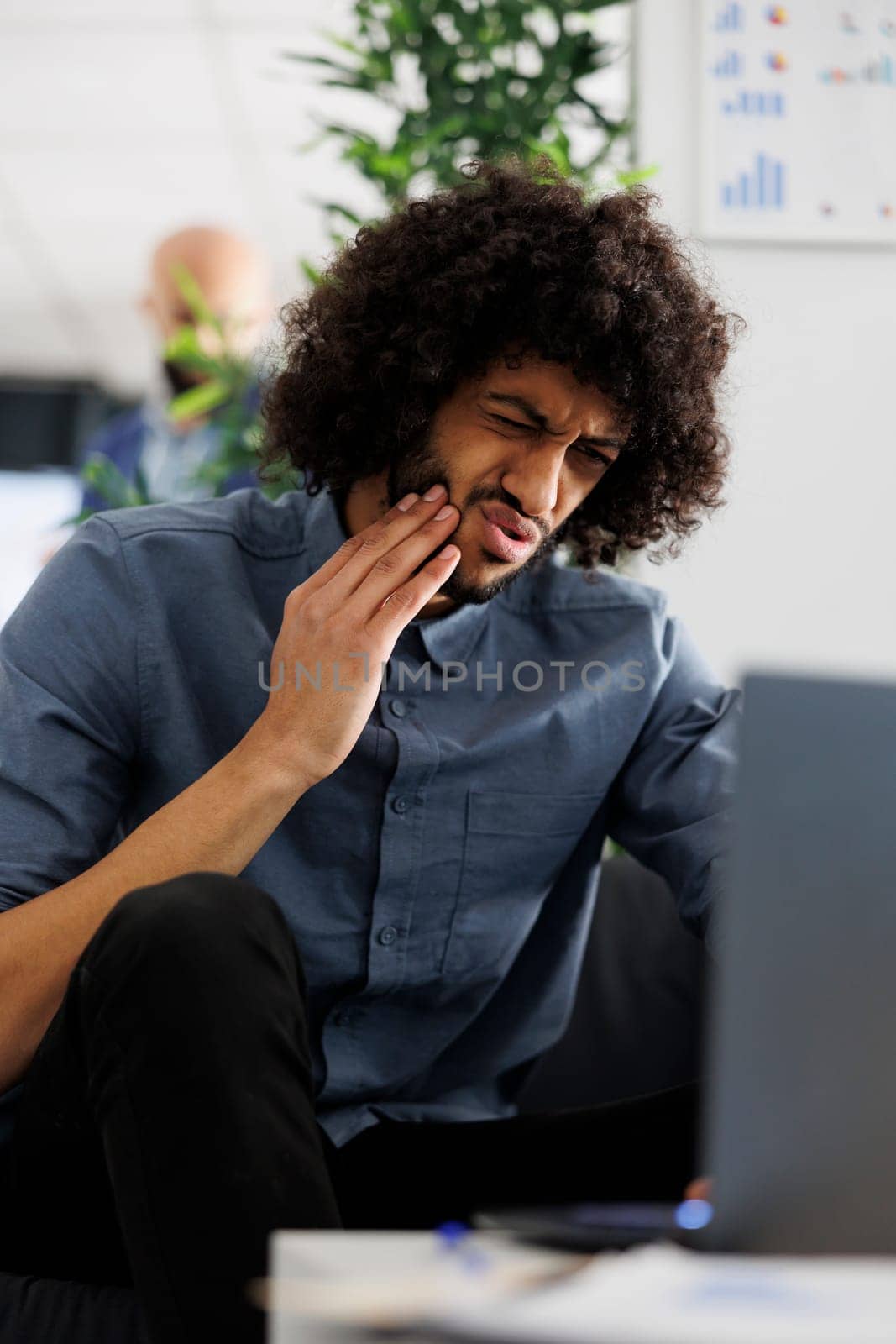 Young arab business employee suffering from toothache at workplace while working on laptop. Entrepreneur having tooth pain while sitting on couch in start up company office