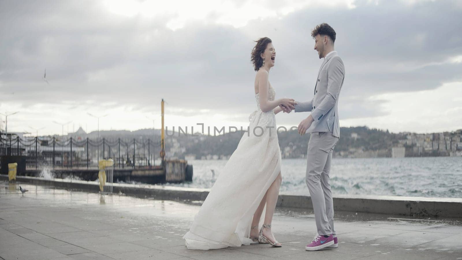 Romantic wedding couple hugging each other and standing on pier against blurry cityscape and green hills. Action. Raging waves and bride and groom laughing out loud