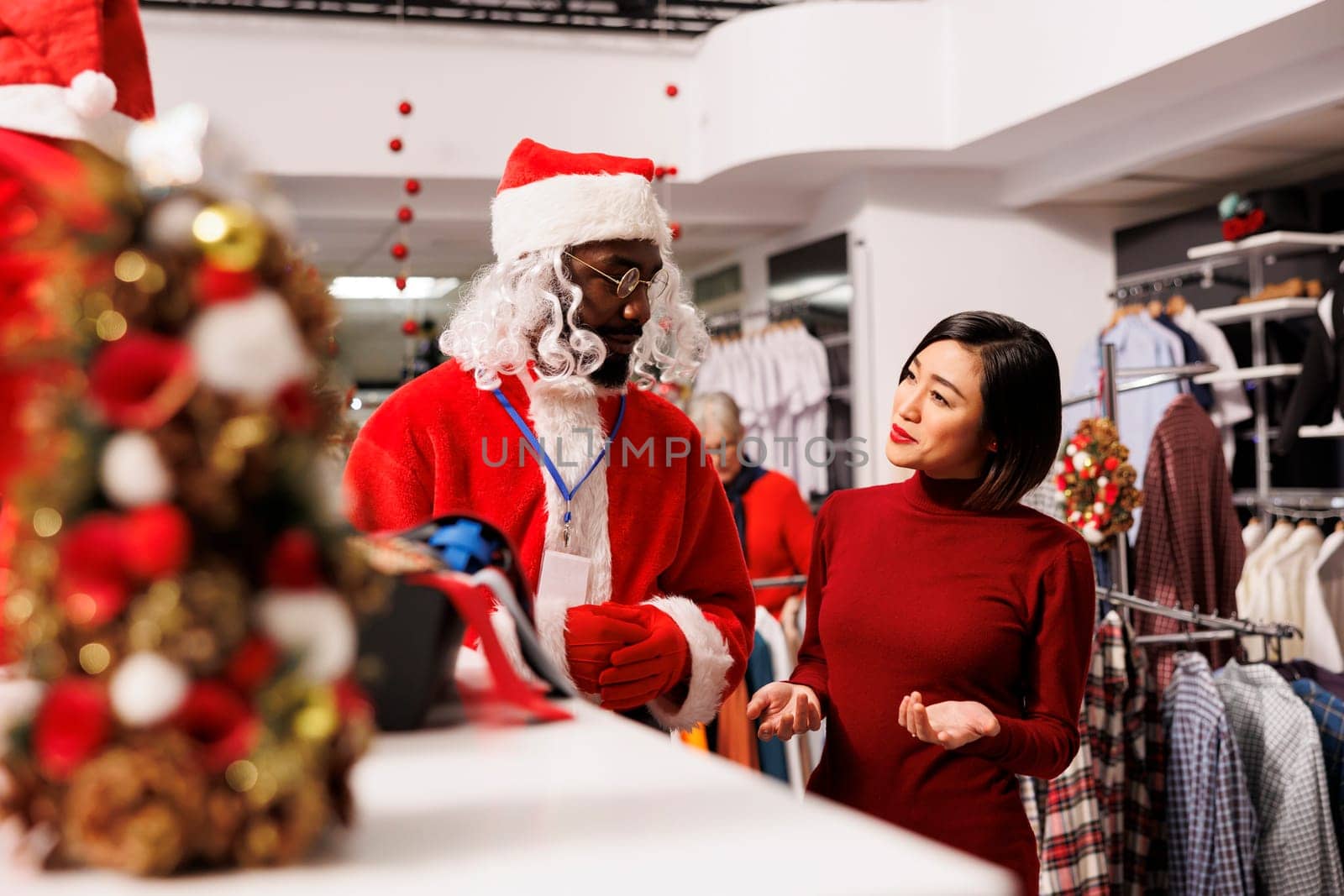 Store worker in santa suit helps client and presenting new clothing collection at shopping mall, talking to asian woman about christmas present ideas. Festive person in costume greeting buyer.