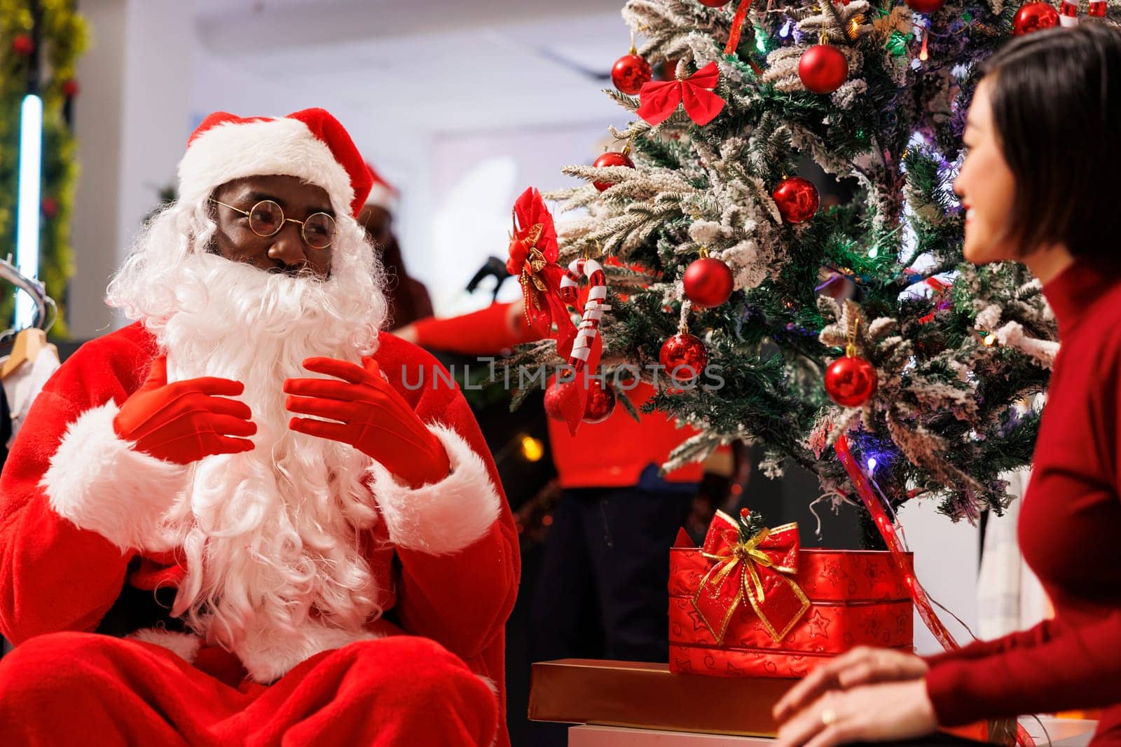 Mall worker Santa Claus sitting next to xmas tree with asian client in festive decorated retail store. Fashion boutique employee offering gift to happy customer, discussing about winter wishes.