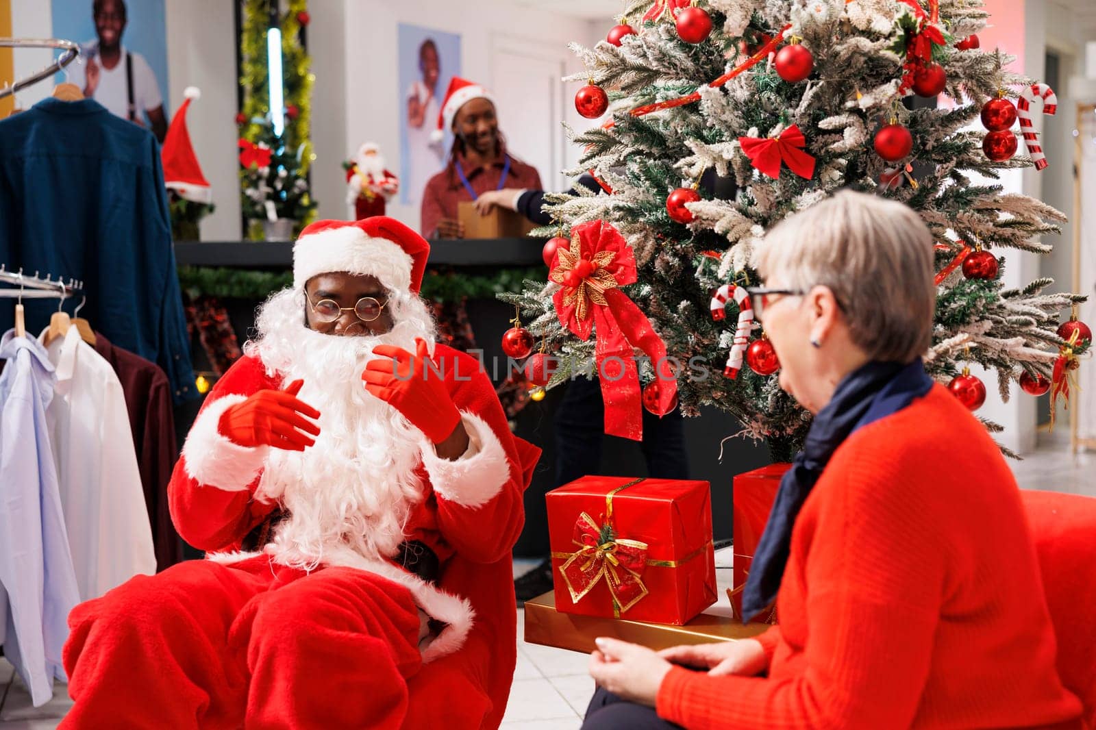 Santa claus employee gives presents to elderly woman in store, offering festive box next to xmas tree at mall. Senior person feeling cheerful after receiving goods for free, seasonal sales.