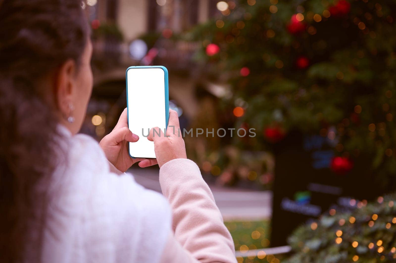 Close-up hands of a woman in winter clothes outdoors next to the Christmas tree, holding smart mobile phone with white blank mockup digital screen, copy ad space. Merry Christmas and New Year concept.