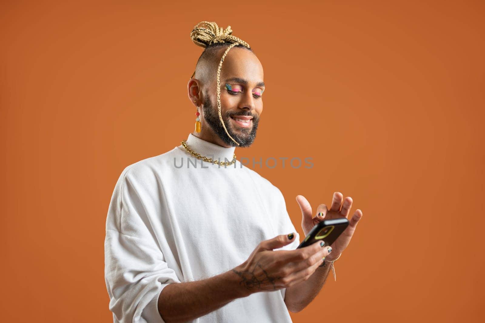 Latino gay man with bright makeup use smartphone isolated orange background. Confident Brazilian gay dressed white using mobile phone, sending message in social media or make online shopping