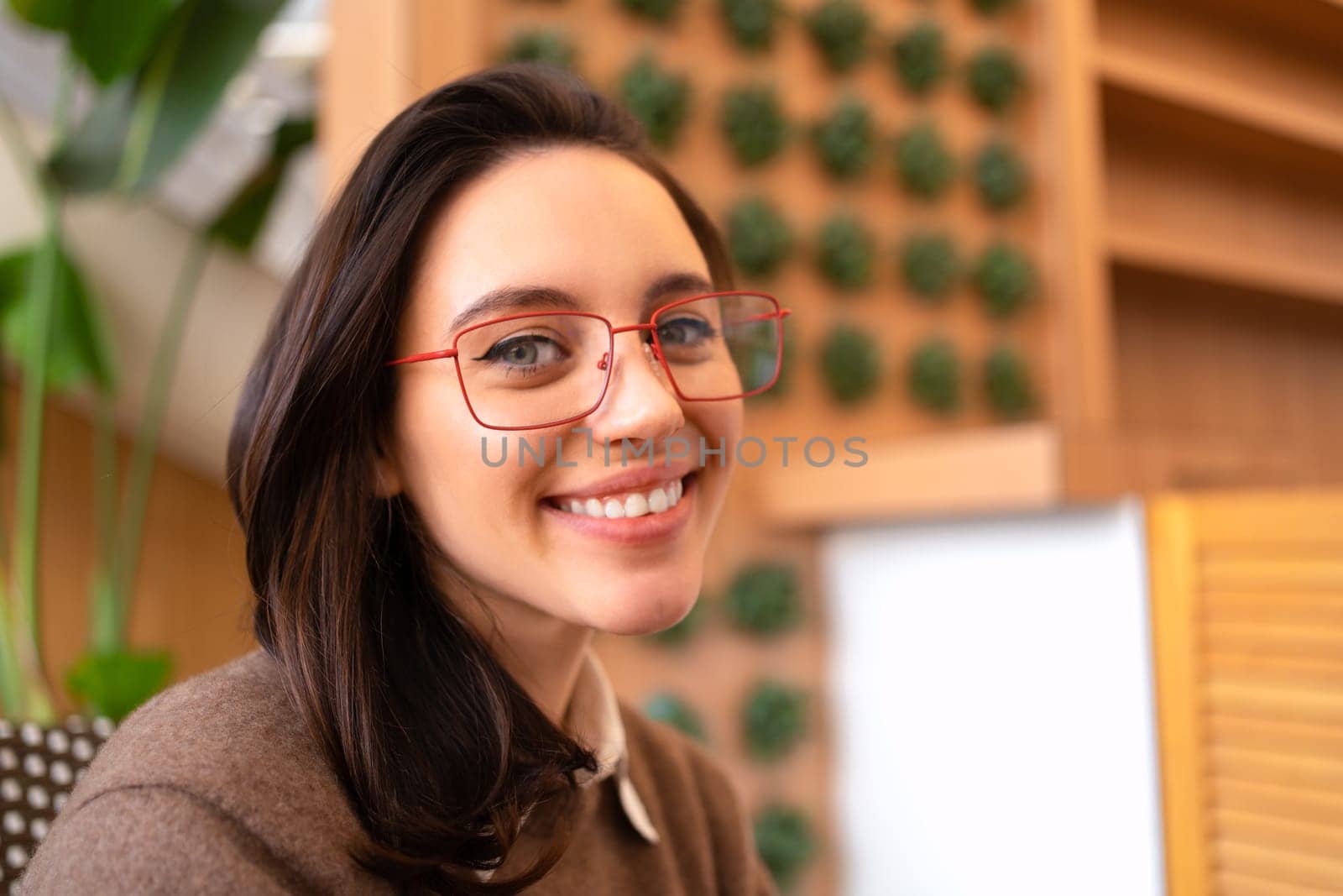 Young adult woman wear glasses smiles looking at camera close up portrait. Casual cheerful woman with eyeglasses smiling at camera on blur background. Close up of happy young female laughing with eyeglasses