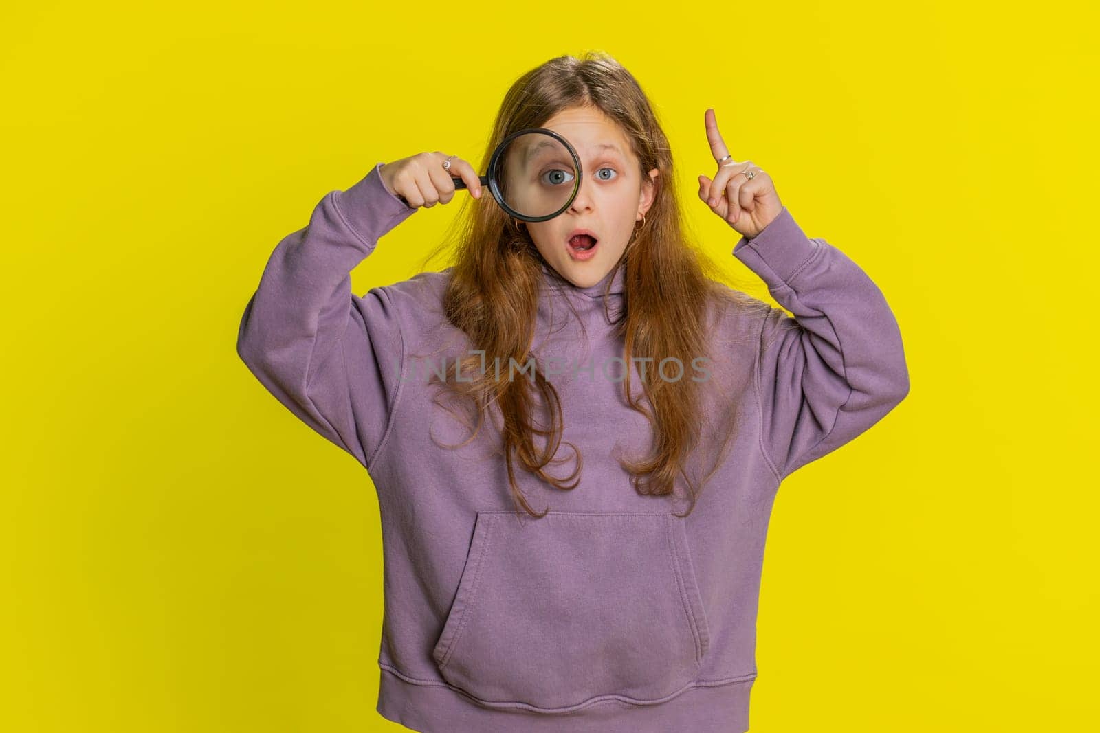 Investigator researcher scientist preteen child girl kid holding magnifying glass near face, looking into camera with big zoomed funny eye searching analyzing inspiration motivation, yellow background