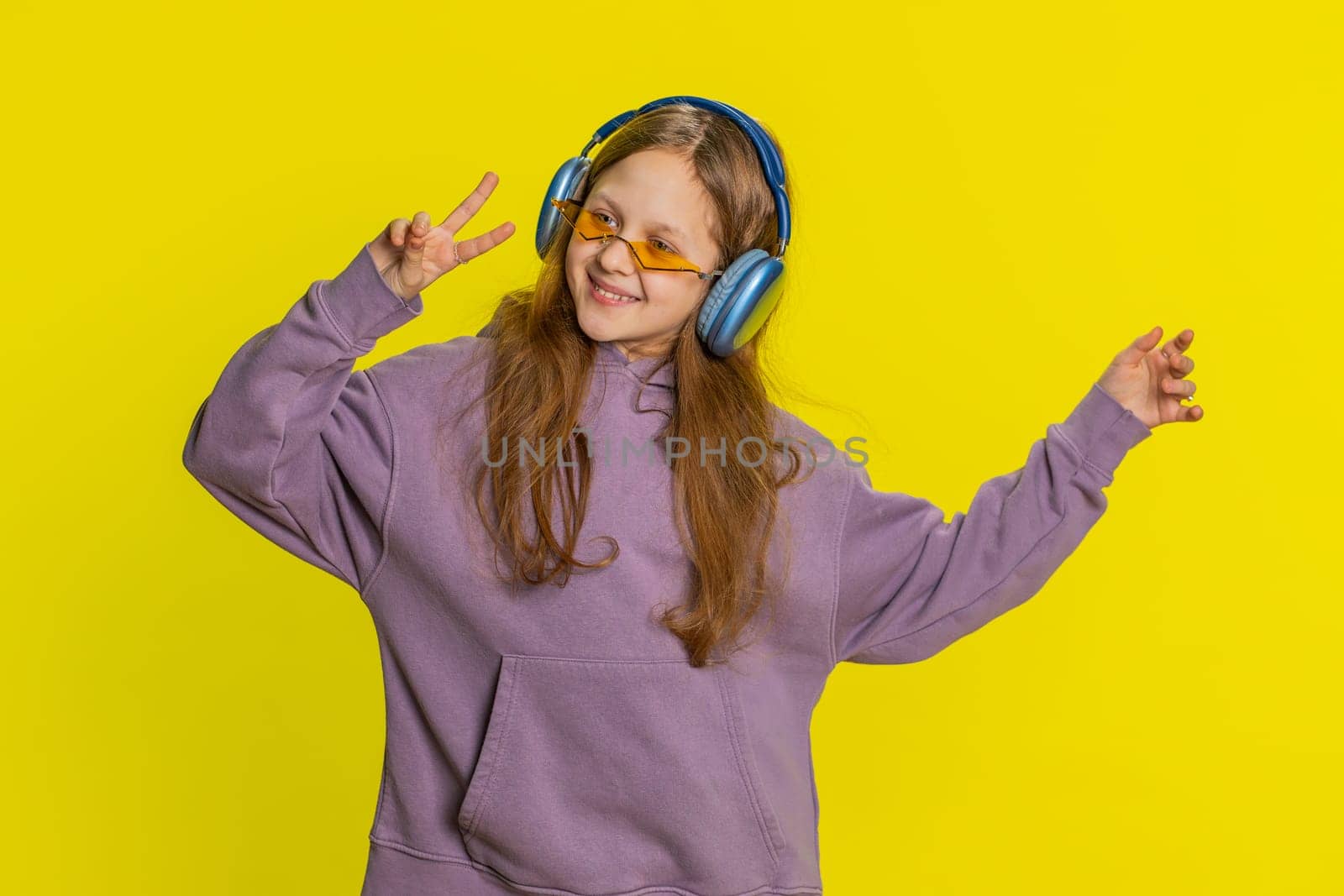 Happy smiling preteen child girl in headphones listening music, dancing disco, fooling around having fun expressive gesticulating hands relaxing on party, making funny moves. Kid on yellow background
