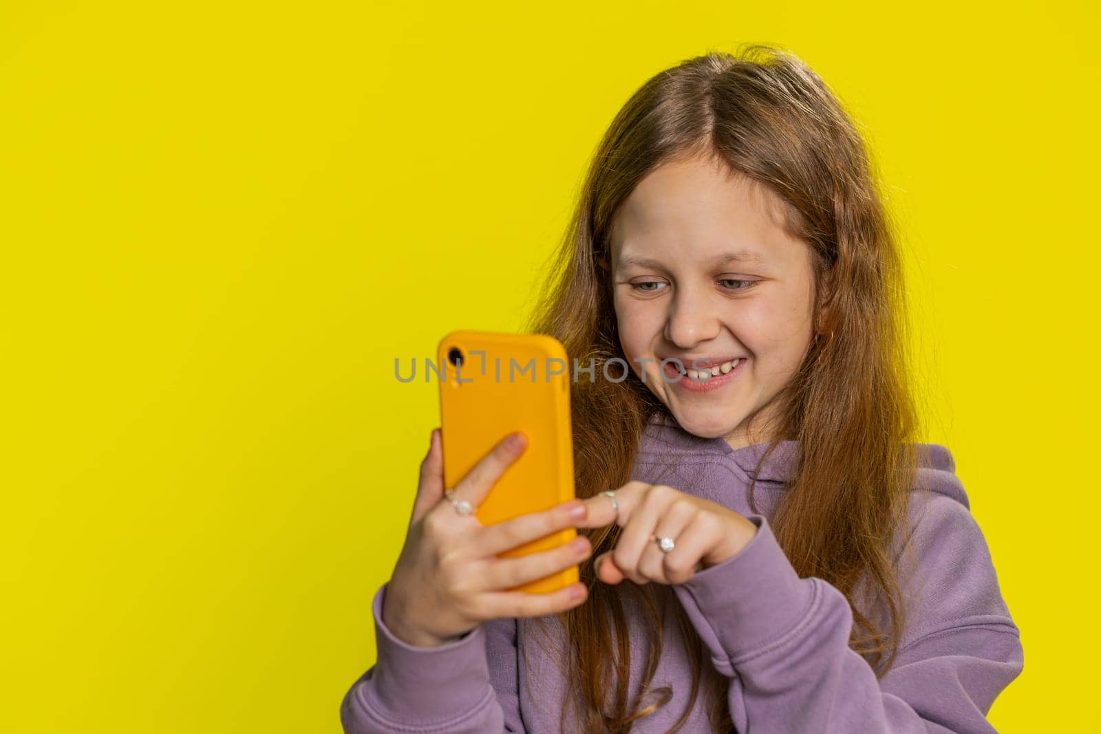 Smiling preteen child girl kid using mobile smartphone typing new post on web, sms message watching video, browsing, addiction of social media networks. Teenager children isolated on yellow background