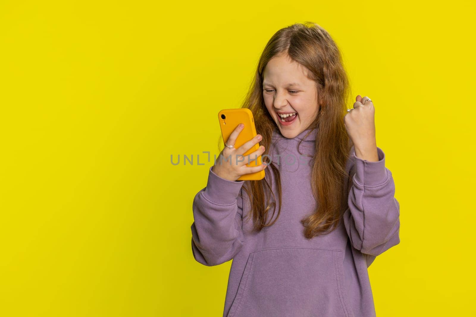 Young preteen child girl kid use mobile smartphone typing browsing say Wow yes found out great big win good news celebrate doing winner gesture. Little cute children on studio yellow background