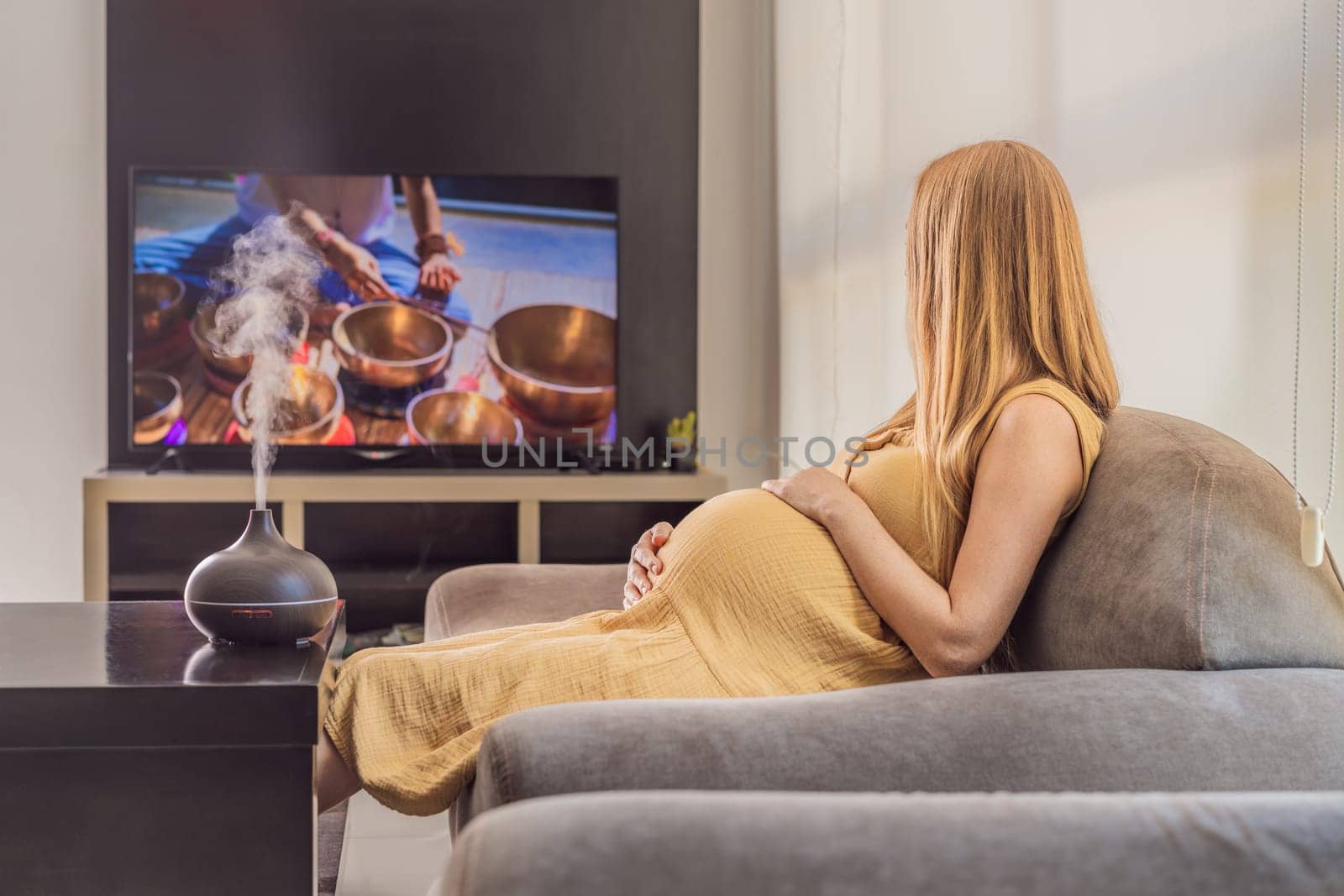 A blissful pregnant woman immerses in relaxation, savoring the soothing aroma from a diffuser while indulging in a calming TV video, embracing tranquility during her pregnant journey by galitskaya