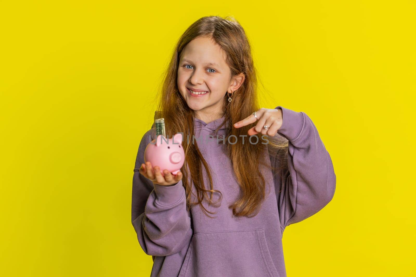 Young preteen child girl kid putting dollar pocket money cash into piggy bank saving for future education. Teenager children with coins box finance income growth, economy on yellow background indoors
