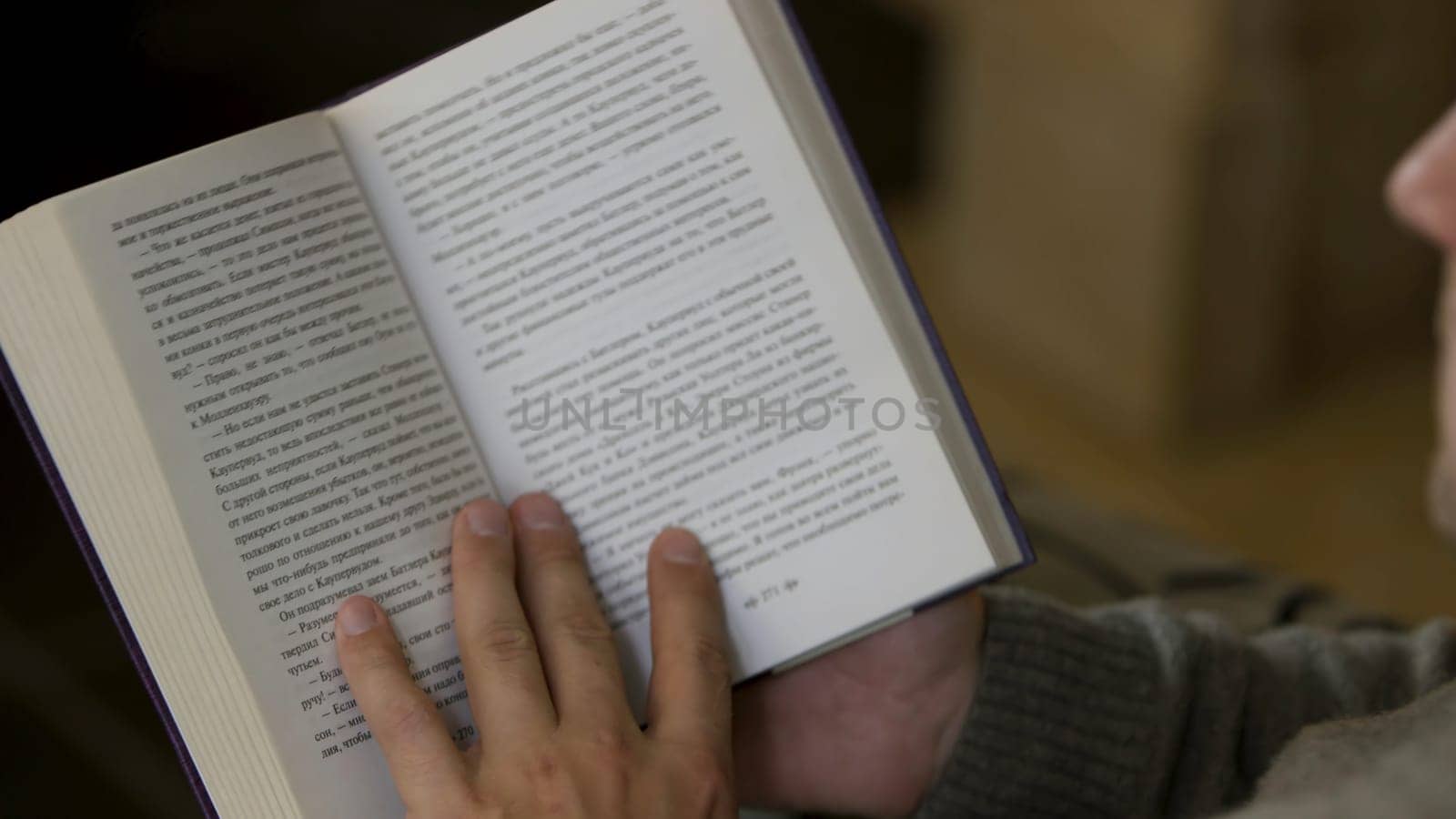 Over the shoulder view of a man reading a book at home. Middle aged male sitting and reading a paper book, turning pages, relaxing at home.