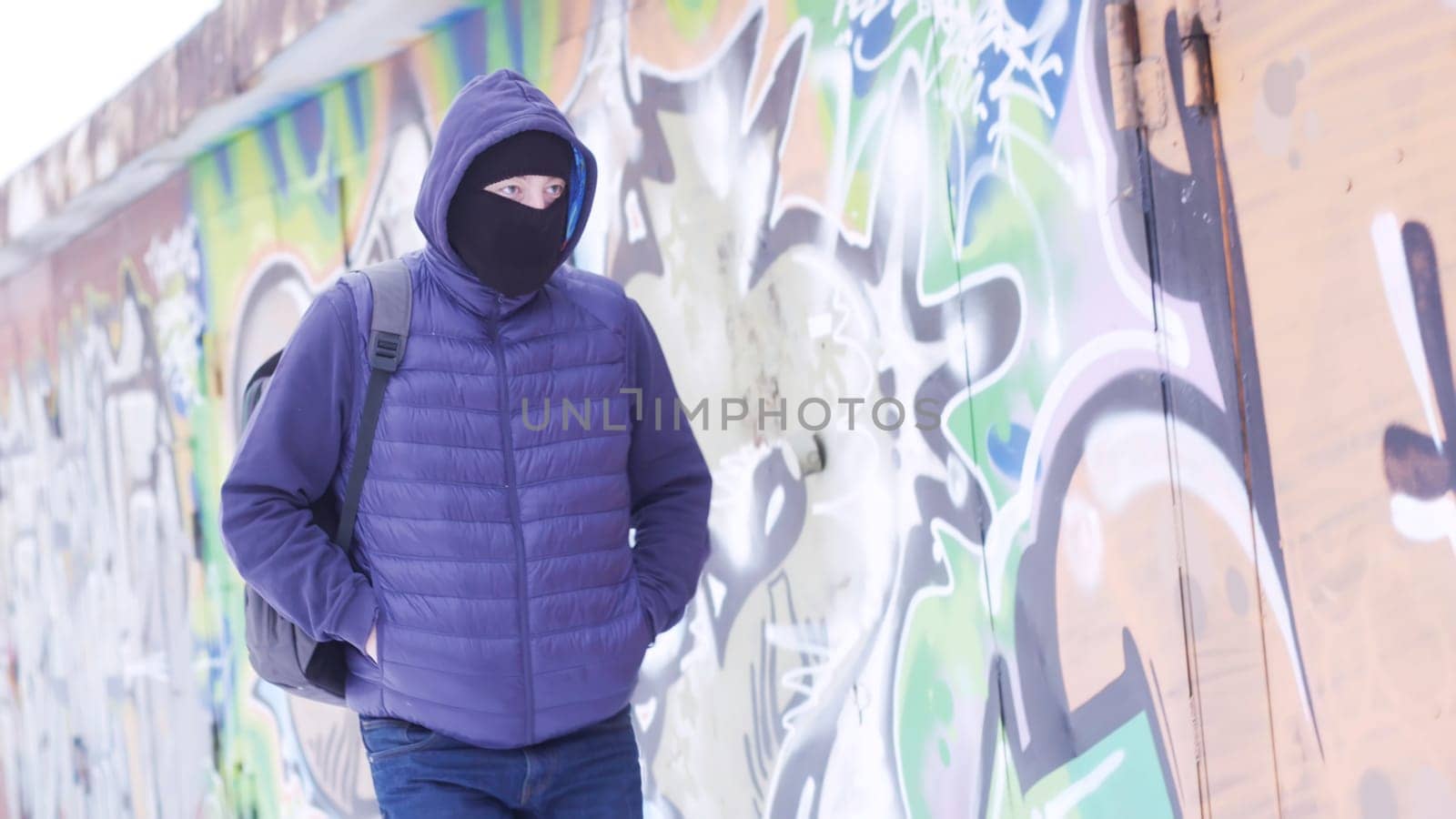 Serious young man in casual outfit and balaclava walking along the grafitti wall. Male with a backpack hiding his face and looking around, concept of vandalism.
