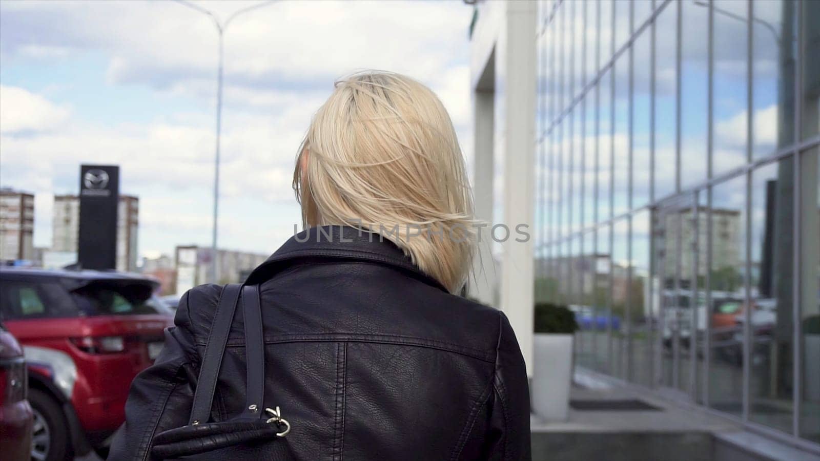 Rear view of a blonde in a black leather jacket walking in the city street. Young, blonde woman walking through the streets of the city with a bag over her shoulder.