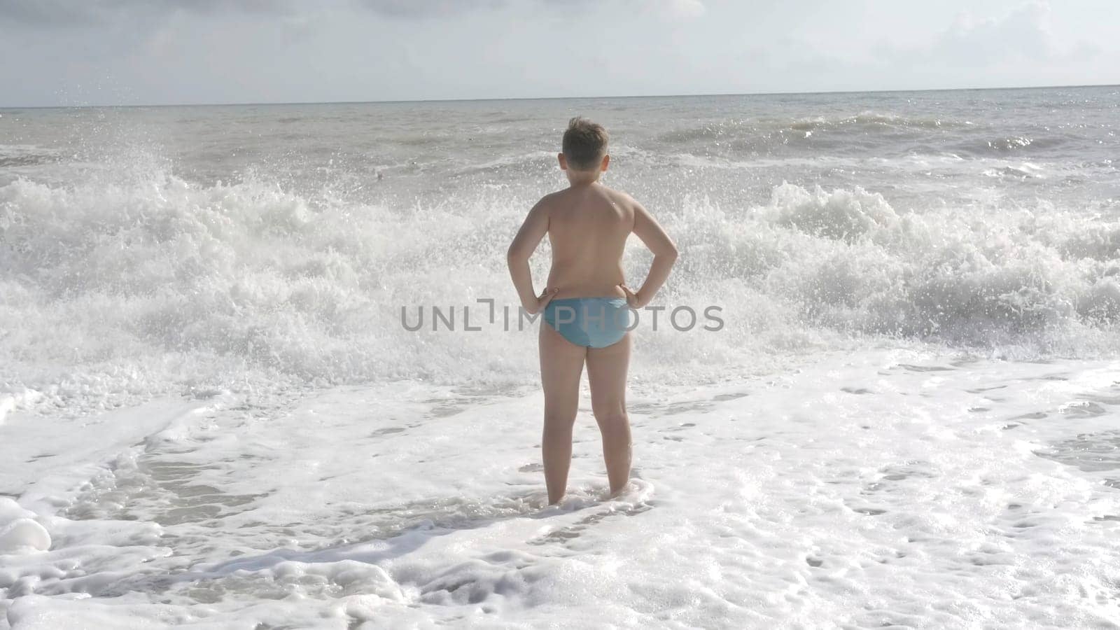 A boy who stands on the waves and admires the view of the sea. Creative . A boy in swimming shorts stands in the sea and looks at the sea waves. by Mediawhalestock