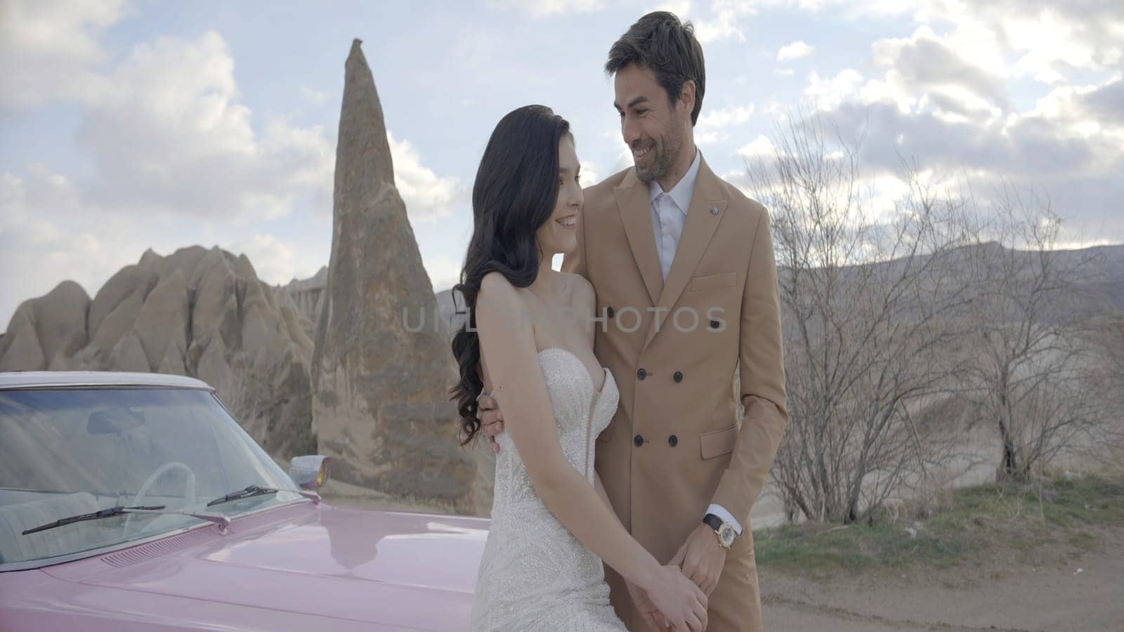 Beautiful couple with car on background of mountains. Action. Elegant outfits of newlyweds vacationing in mountains. Stylish newlyweds sit on hood of car on background of canyon.